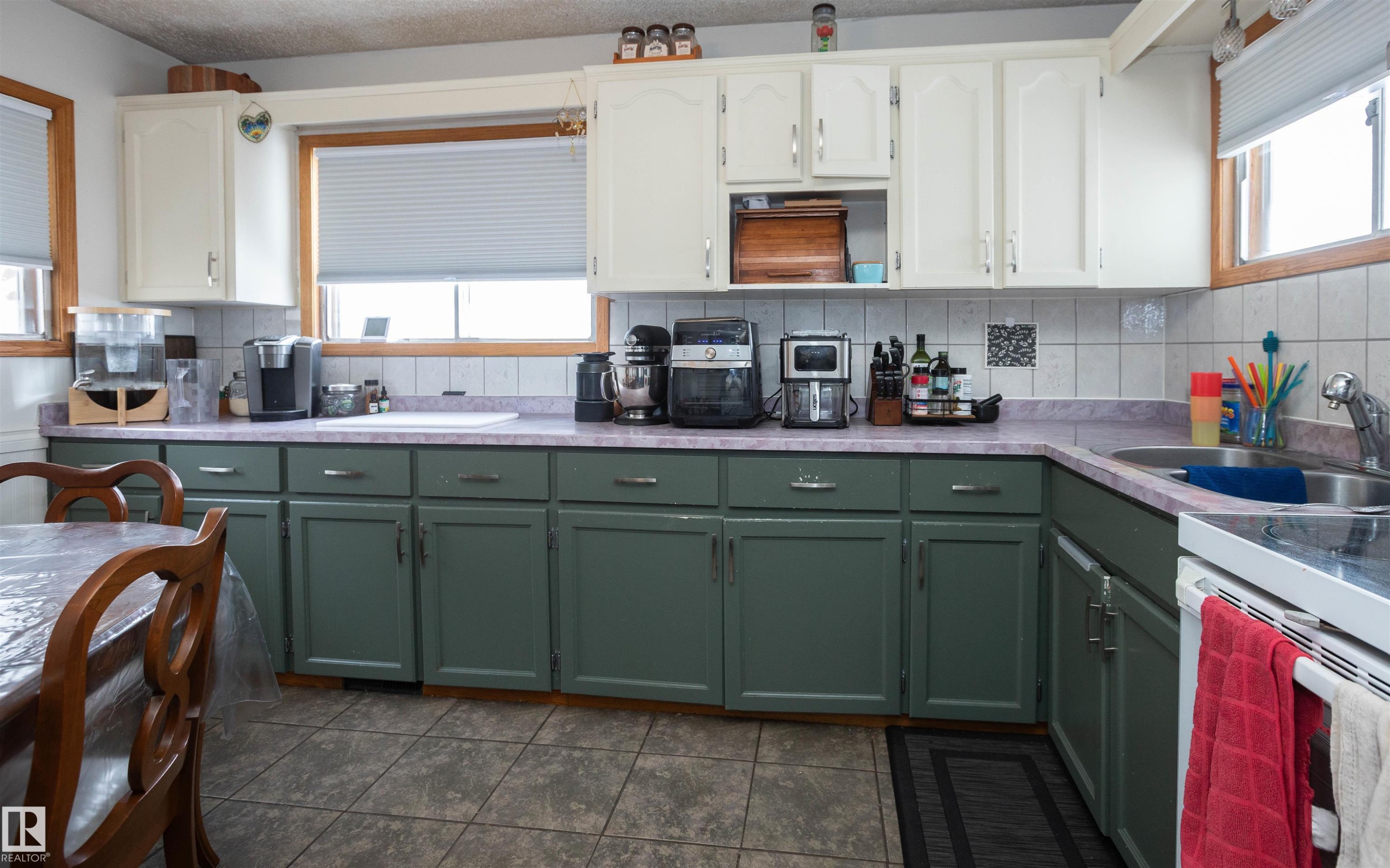 The kitchen features white upper cabinetry and green lower cabinetry, a light-colored tile backsplash, and a double basin stainless steel sink - 4420 56 Avenue, Barrhead, AB - Indoor Photo Showing Kitchen With Double Sink