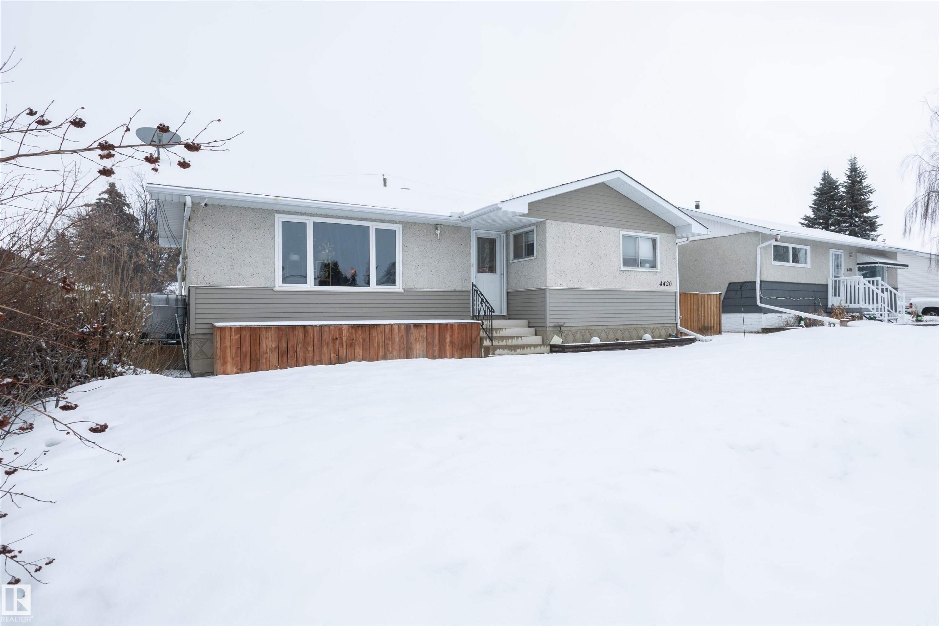 The property features a light-colored stucco exterior with contrasting siding, a prominent front window, and a white front door accessible by exterior steps - 4420 56 Avenue, Barrhead, AB - Outdoor