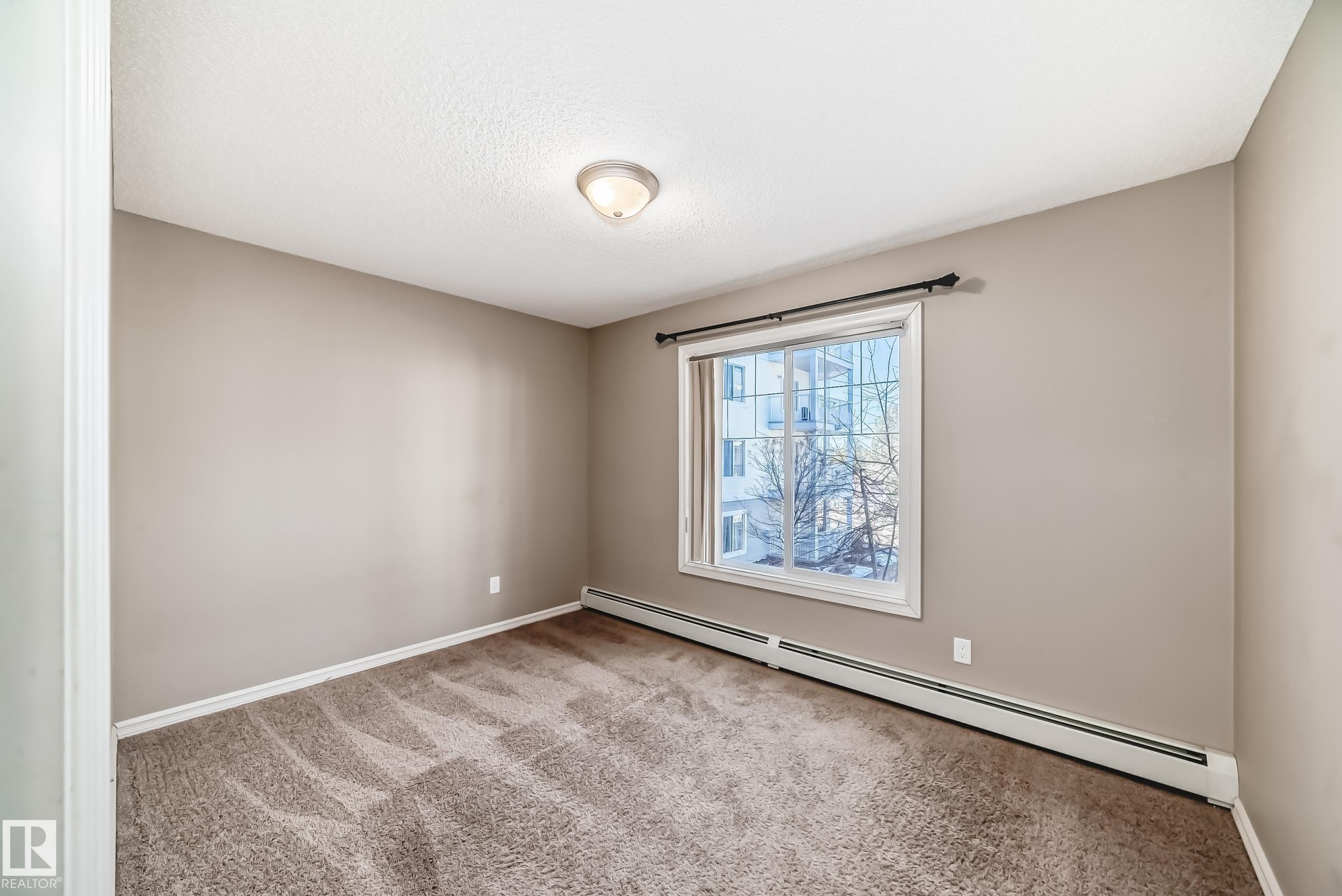 This room features neutral-toned walls, a textured carpet, and a window providing natural light and exterior views - 208 2204 44 Ave, Edmonton, AB - Indoor Photo Showing Other Room