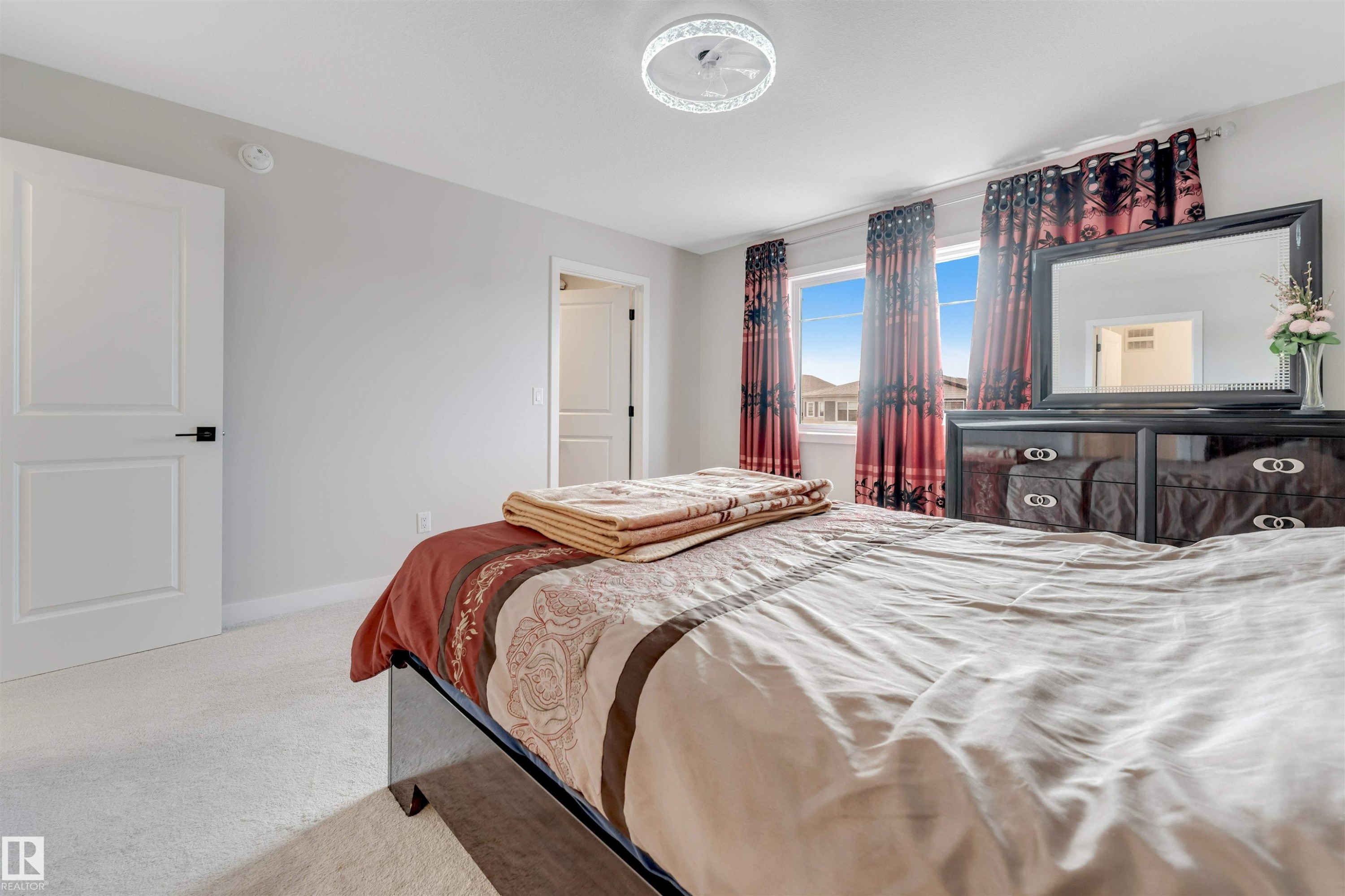 This bedroom features carpeted flooring, a ceiling light fixture, a window with red and black patterned curtains, and light gray walls - 6404 27 Avenue, Edmonton, AB - Indoor Photo Showing Bedroom