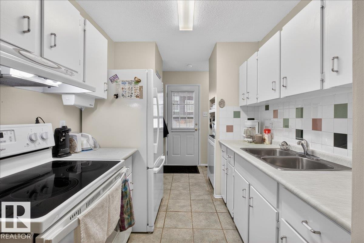 The kitchen features white cabinetry, a dual basin stainless steel sink, a tile backsplash with accent tiles, and a white refrigerator - 2 Mcleod Place, Edmonton, AB - Indoor Photo Showing Kitchen With Double Sink
