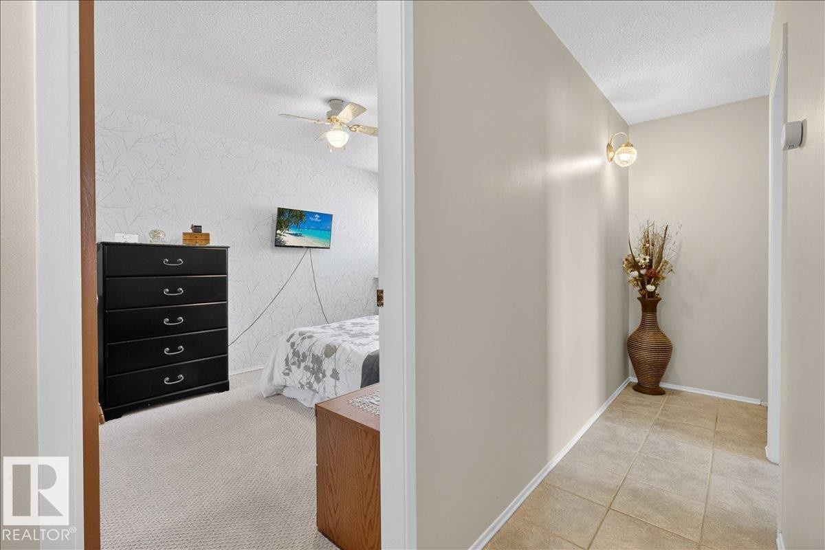 The property features a hallway with tiled flooring and a wall-mounted light fixture, leading to a room with carpeted flooring and a ceiling fan - 2 Mcleod Place, Edmonton, AB - Indoor