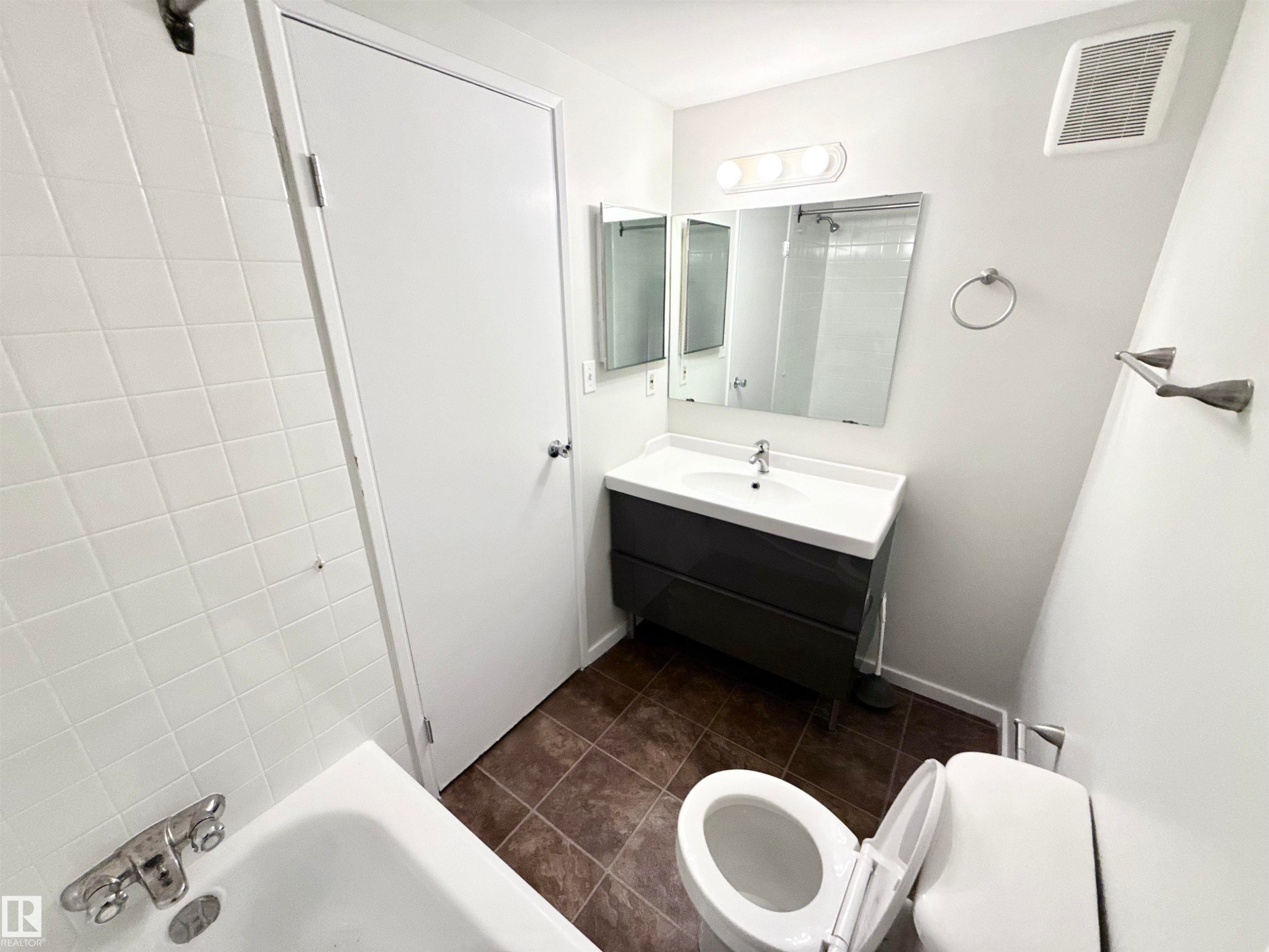 The bathroom features a vanity with a rectangular sink, a toilet, a bathtub with white tiled walls, and a wall-mounted mirror - 307 1620 48 Street, Edmonton, AB