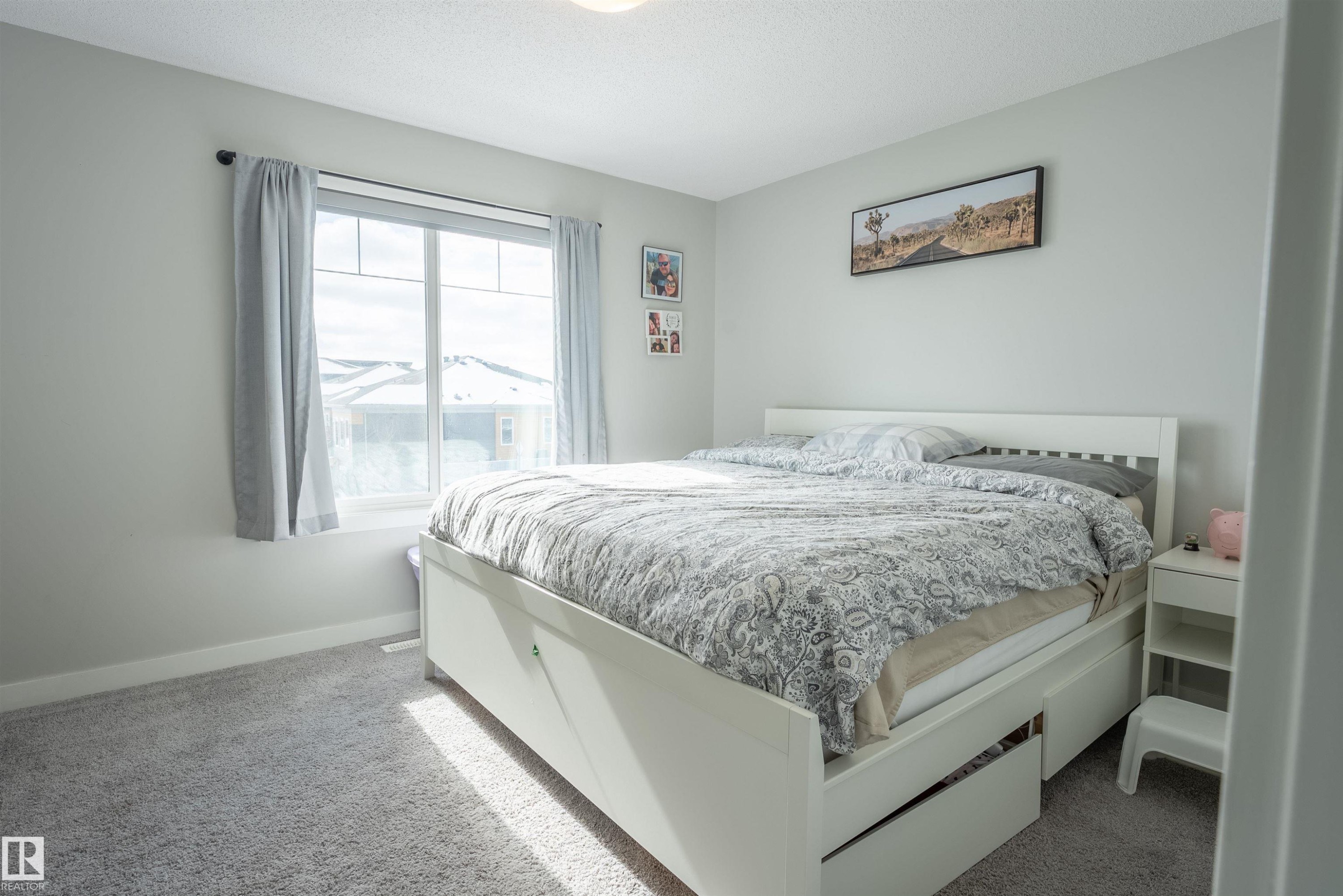 The property features a room with light grey walls and grey carpet, a white bed frame with integrated storage drawers, and a window with light grey curtains - 656 Black Stone, Leduc, AB - Indoor Photo Showing Bedroom