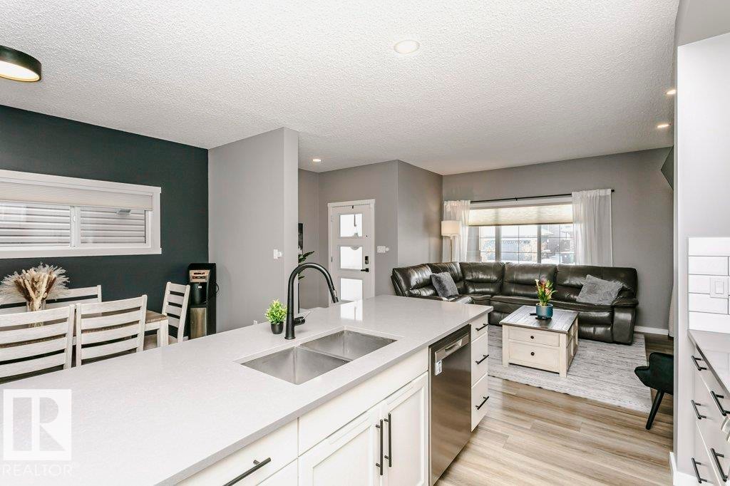 Open concept living space featuring a kitchen island with a double sink, a dining area with a window, and a living area with a large window - 320 Balsam Link, Leduc, AB - Indoor Photo Showing Kitchen With Double Sink