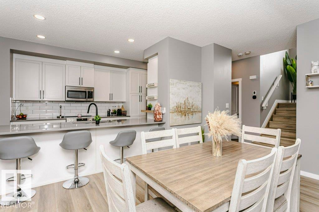 This open-concept living space features light wood-style flooring, a kitchen with white cabinetry and a white subway tile backsplash, and a dining area with a light wood table and white chairs - 320 Balsam Link, Leduc, AB - Indoor