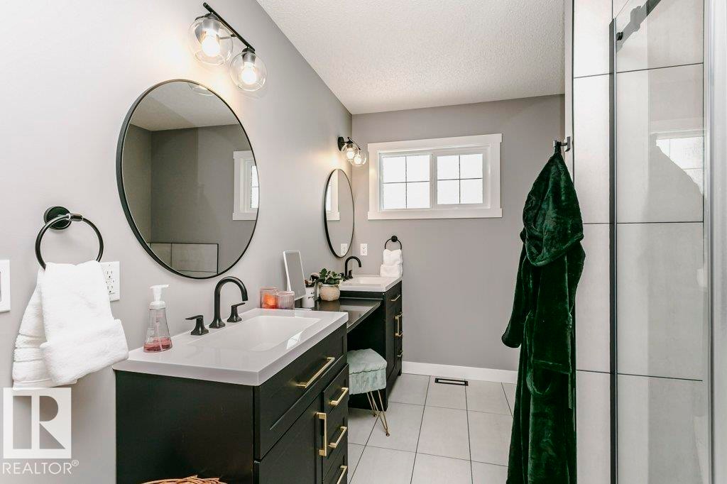 Bathroom featuring a double vanity with dark cabinetry, light countertops, and round mirrors, along with tiled flooring and a window - 320 Balsam Link, Leduc, AB - Indoor Photo Showing Bathroom