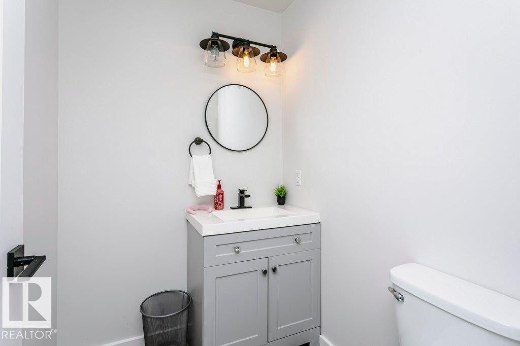 Bathroom featuring a vanity with a white countertop and gray cabinetry, a wall-mounted circular mirror, and a three-light fixture with glass shades - 320 Balsam Link, Leduc, AB - Indoor Photo Showing Bathroom