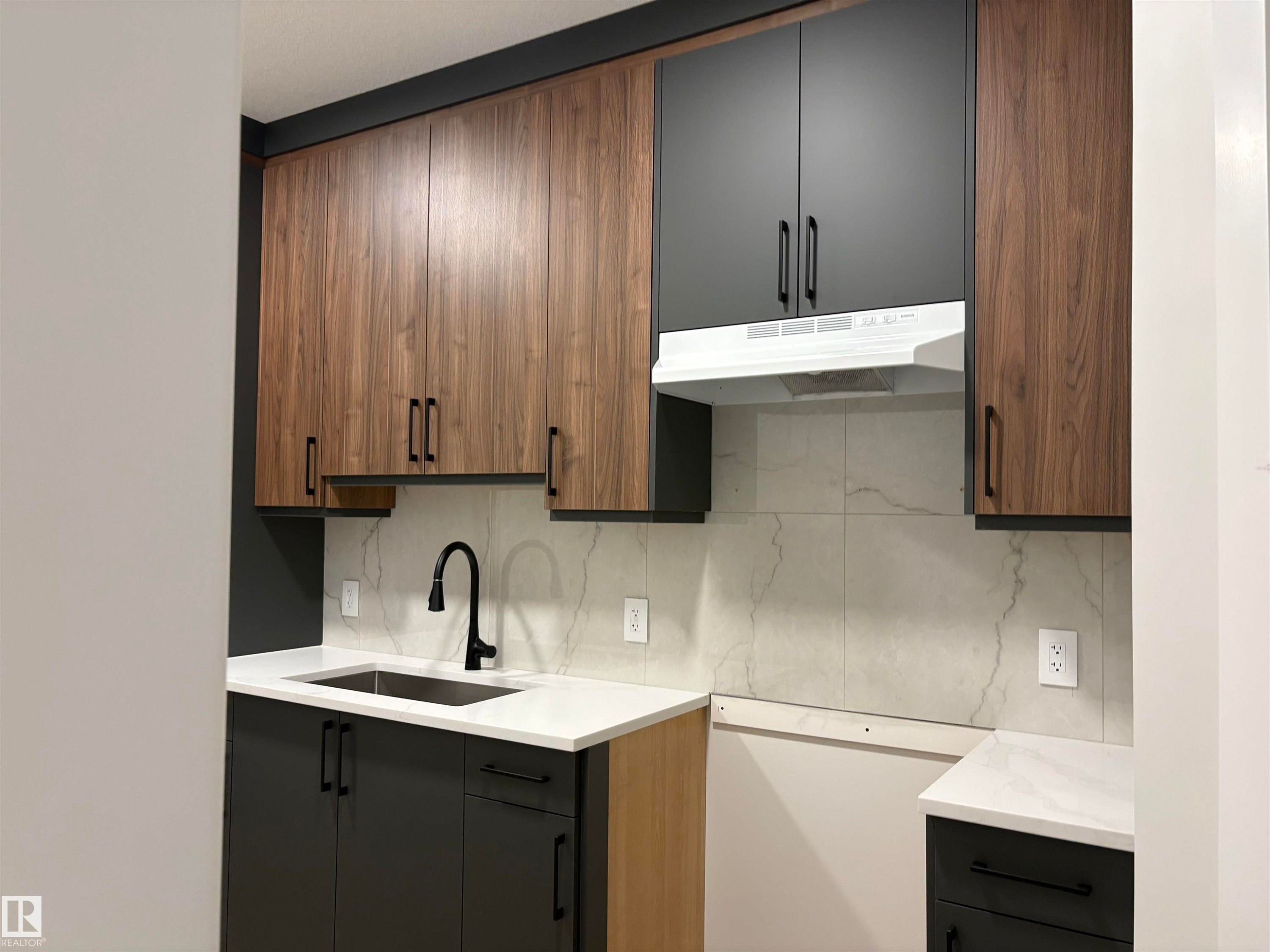 Kitchen featuring wood-finish and matte black cabinetry, light-toned countertops, a matte black gooseneck faucet, and a stone-look tile backsplash - 1039 151 Ave, Edmonton, AB