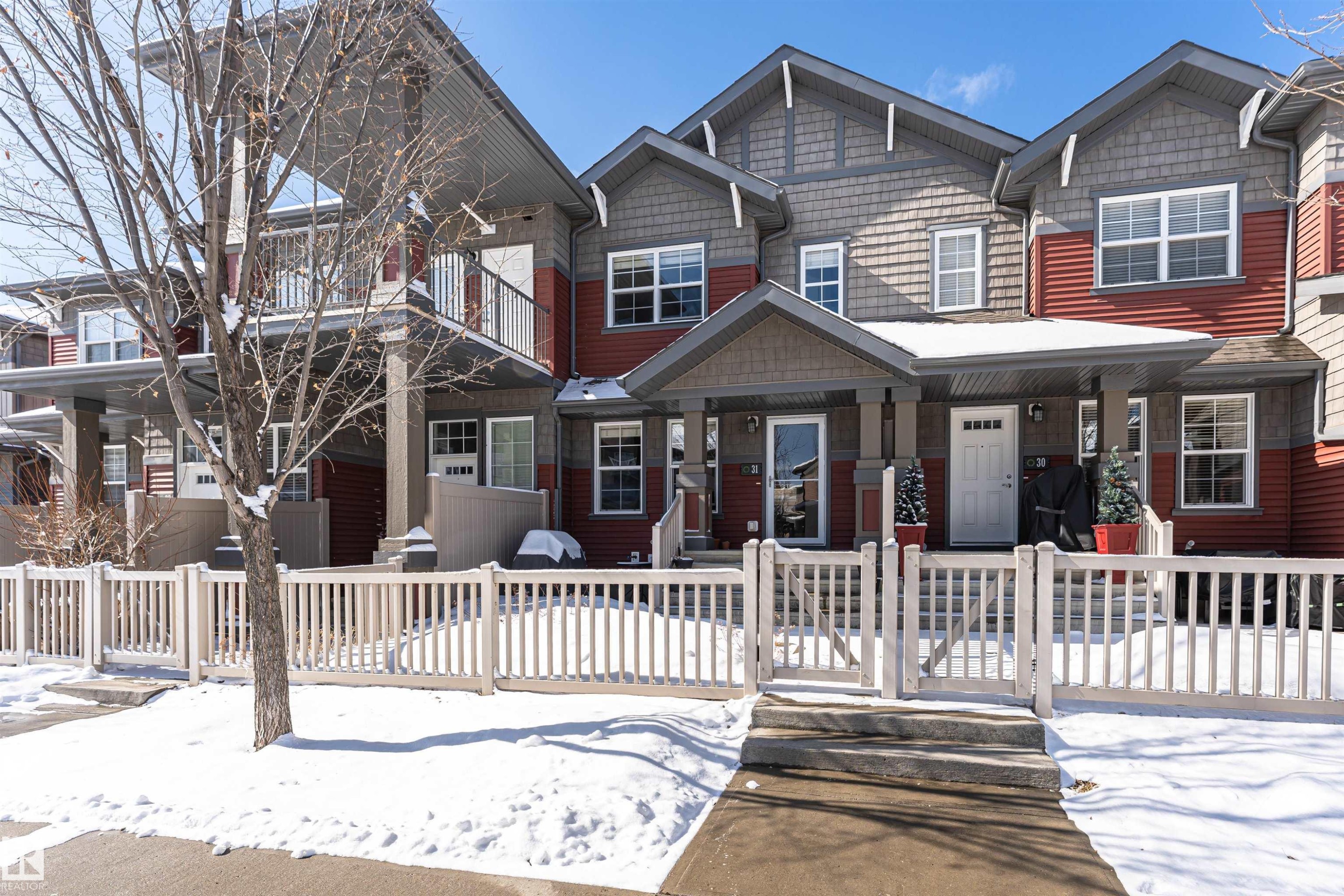 The property presents a facade featuring red and grey siding, white-framed windows, and a white picket fence - 31 4029 Orchards Drive, Edmonton, autreother - Outdoor With Facade