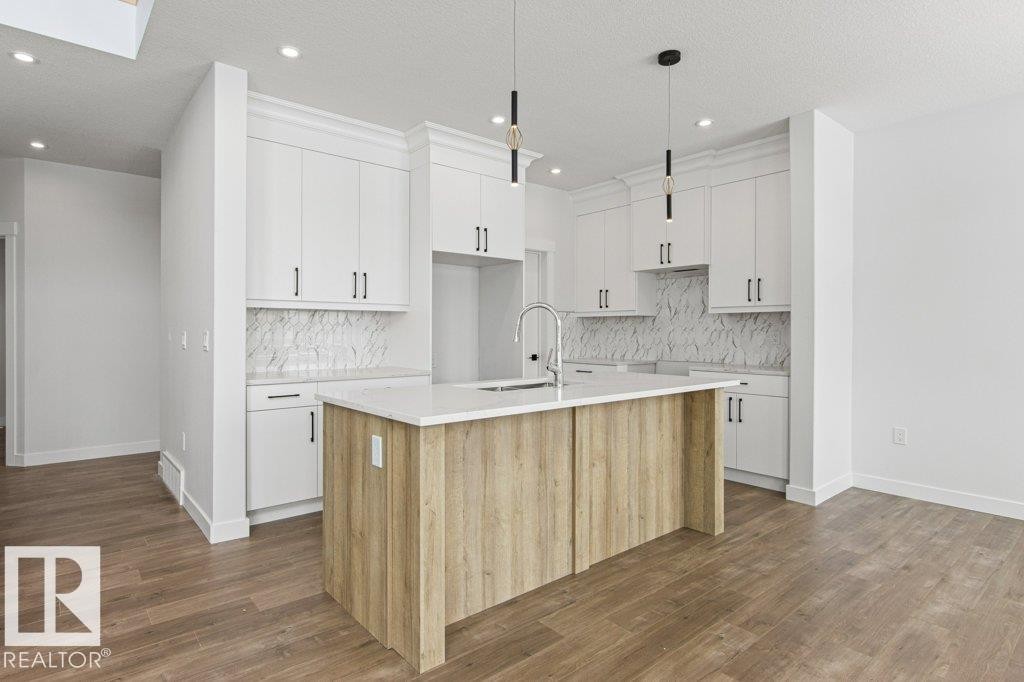 The kitchen features white cabinetry, a light-colored kitchen island with a sink, pendant lighting, and a white backsplash with a patterned design - 5018 53 Avenue, Calmar, AB - Indoor Photo Showing Kitchen With Upgraded Kitchen