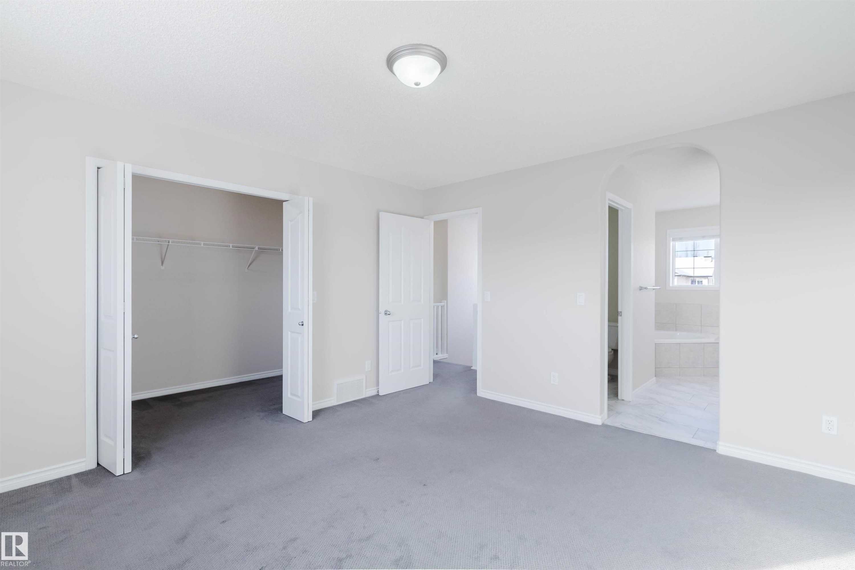 This room features soft gray carpeting, a closet with bi-fold doors and wire shelving, and an arched entryway leading to a bathroom with tiled flooring and a window - 1135 112 Street, Edmonton, AB - Indoor Photo Showing Other Room