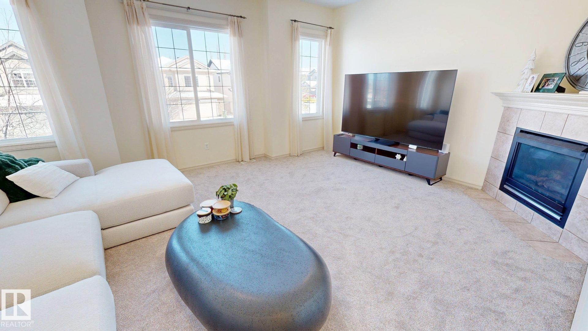 This inviting living space features light-colored carpeting, multiple windows providing natural light, and a fireplace with a tiled surround - 1609 Hodgson Crest, Edmonton, AB - Indoor Photo Showing Living Room With Fireplace