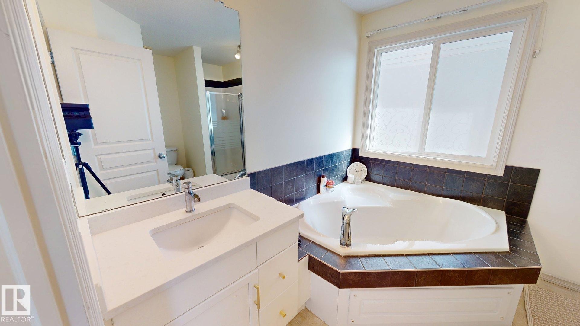 The bathroom features a corner bathtub with dark tiled surround, a window, and a vanity with a white countertop and sink - 1609 Hodgson Crest, Edmonton, AB - Indoor Photo Showing Bathroom