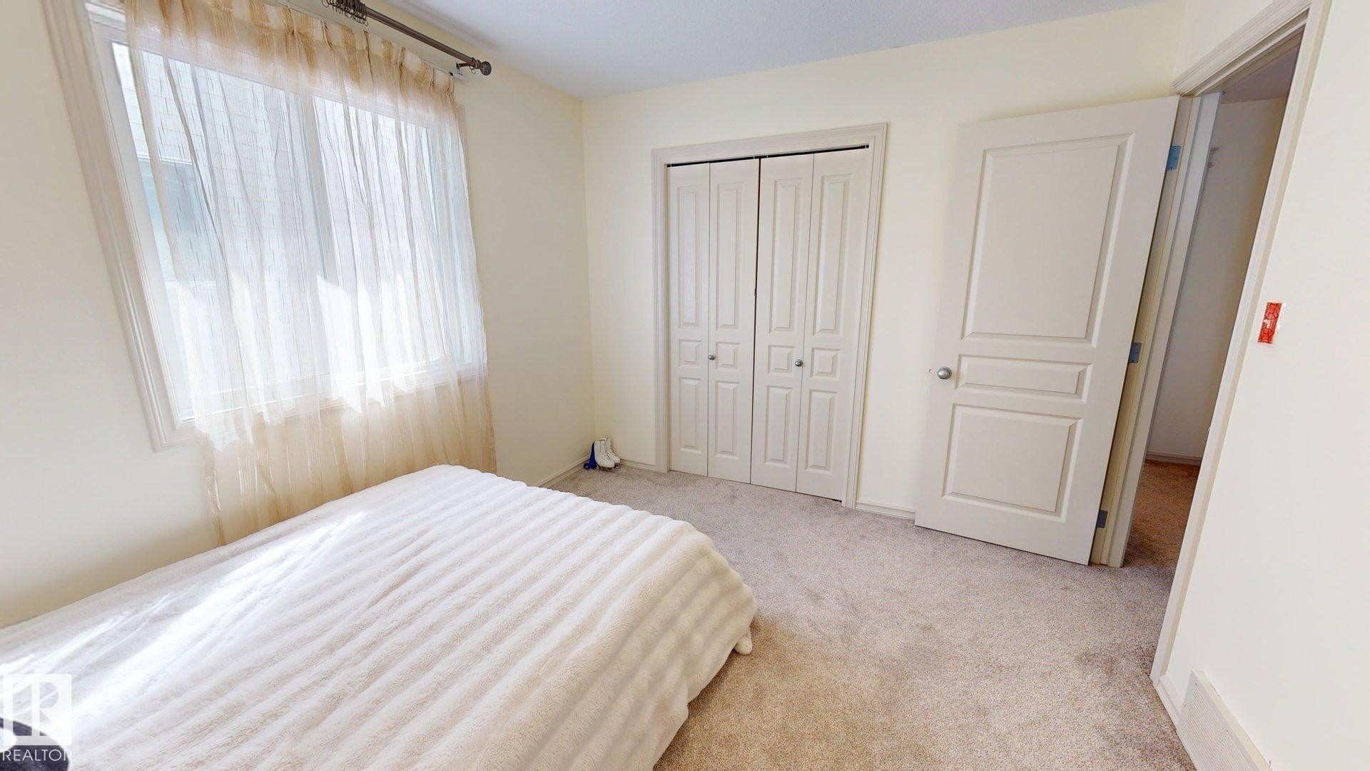 This bright room features a window with sheer curtains, a closet with bi-fold doors, and neutral carpeting - 1609 Hodgson Crest, Edmonton, AB - Indoor Photo Showing Bedroom