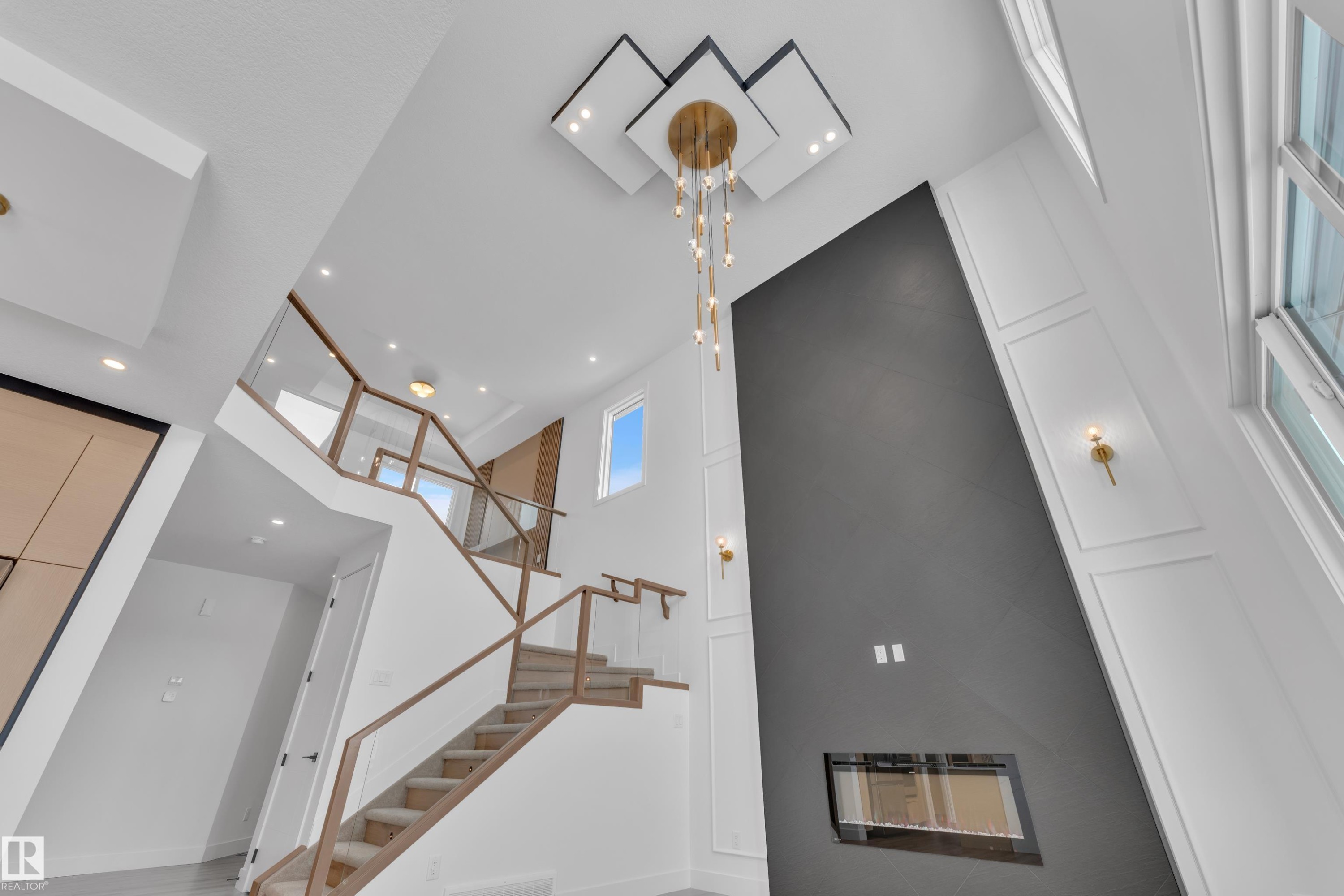 The property features a grand staircase with glass railings and wood handrails, an accent wall with a modern fireplace, and a striking chandelier - 503 Creekside Bay, Leduc, AB - Indoor Photo Showing Other Room