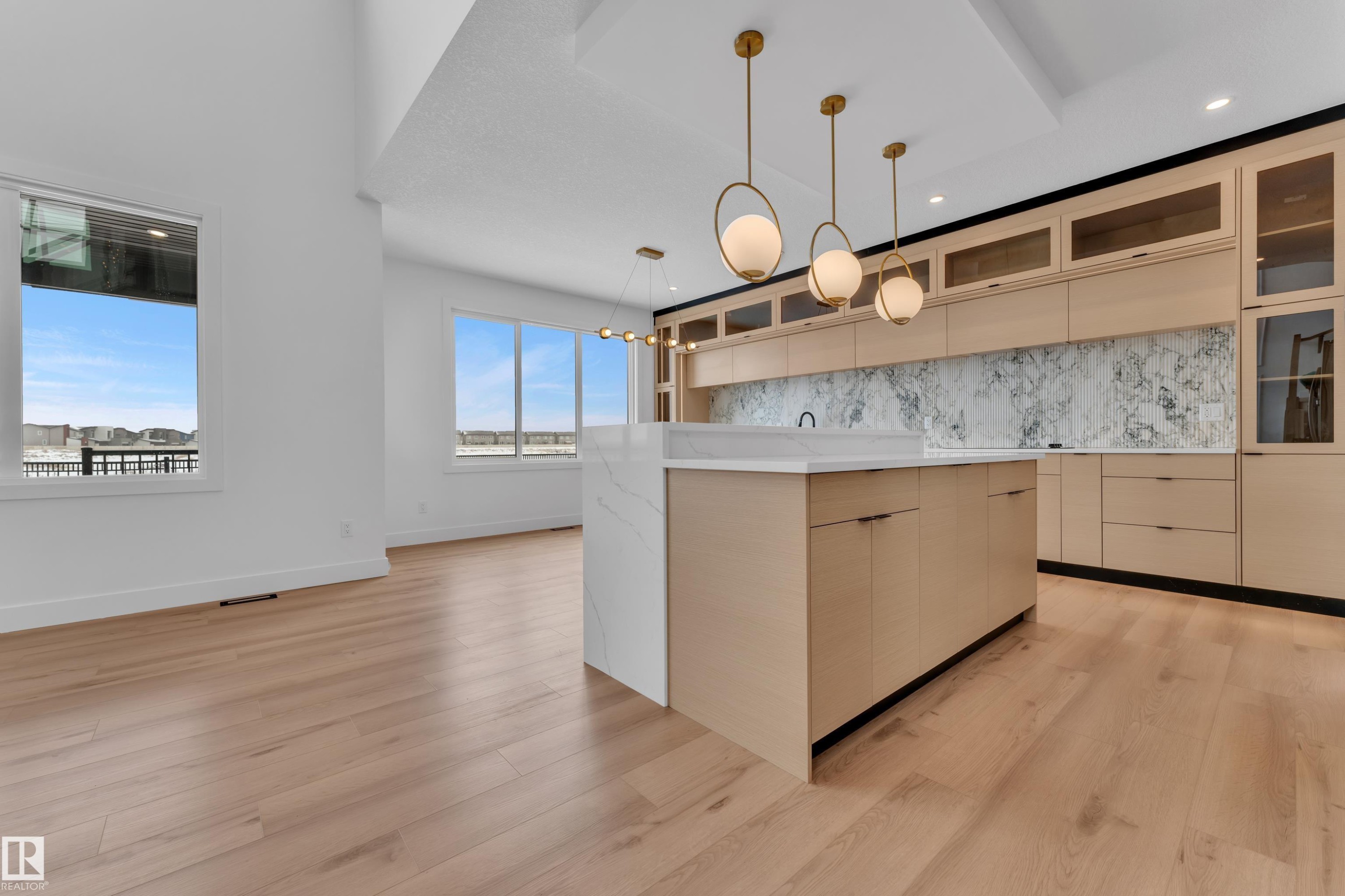 Open concept living space featuring light hardwood flooring, a contemporary kitchen island with a white countertop and waterfall edge, and custom cabinetry with glass-fronted upper cabinets - 503 Creekside Bay, Leduc, AB - Indoor Photo Showing Kitchen