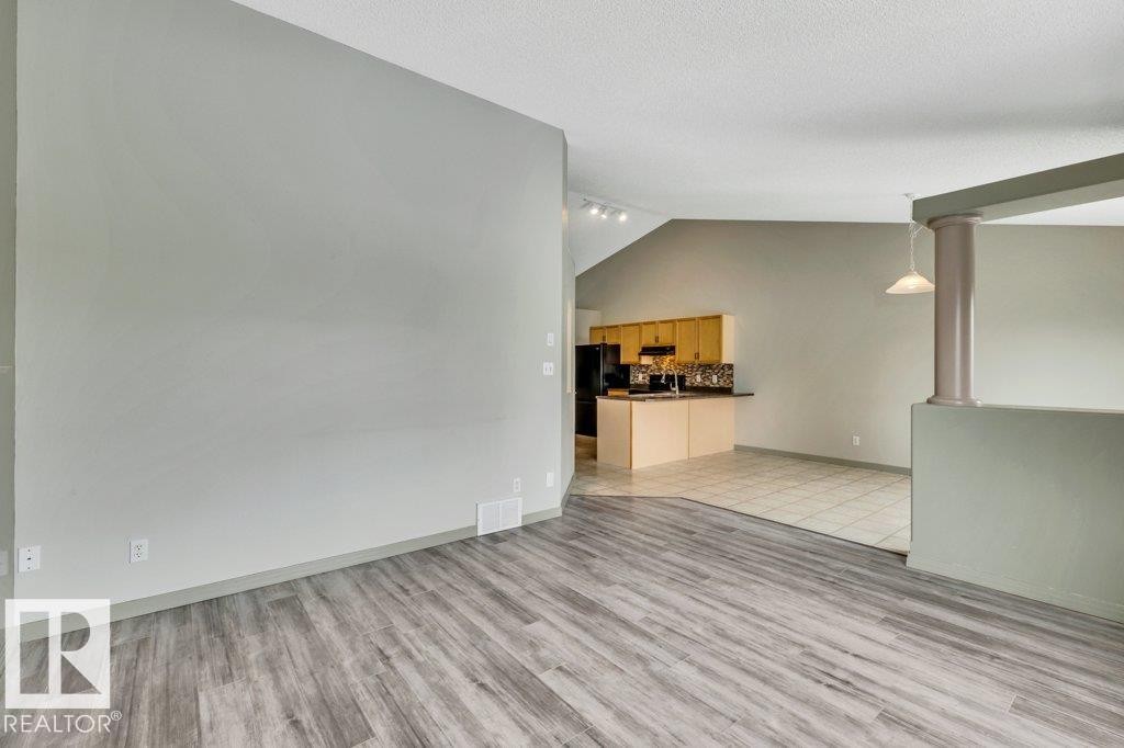 The property features an open-concept layout with wood-look flooring and light-toned walls, leading into a kitchen area with tiled flooring, light wood cabinetry, and a black refrigerator - 902 Graham Wynd, Edmonton, AB - Indoor Photo Showing Other Room