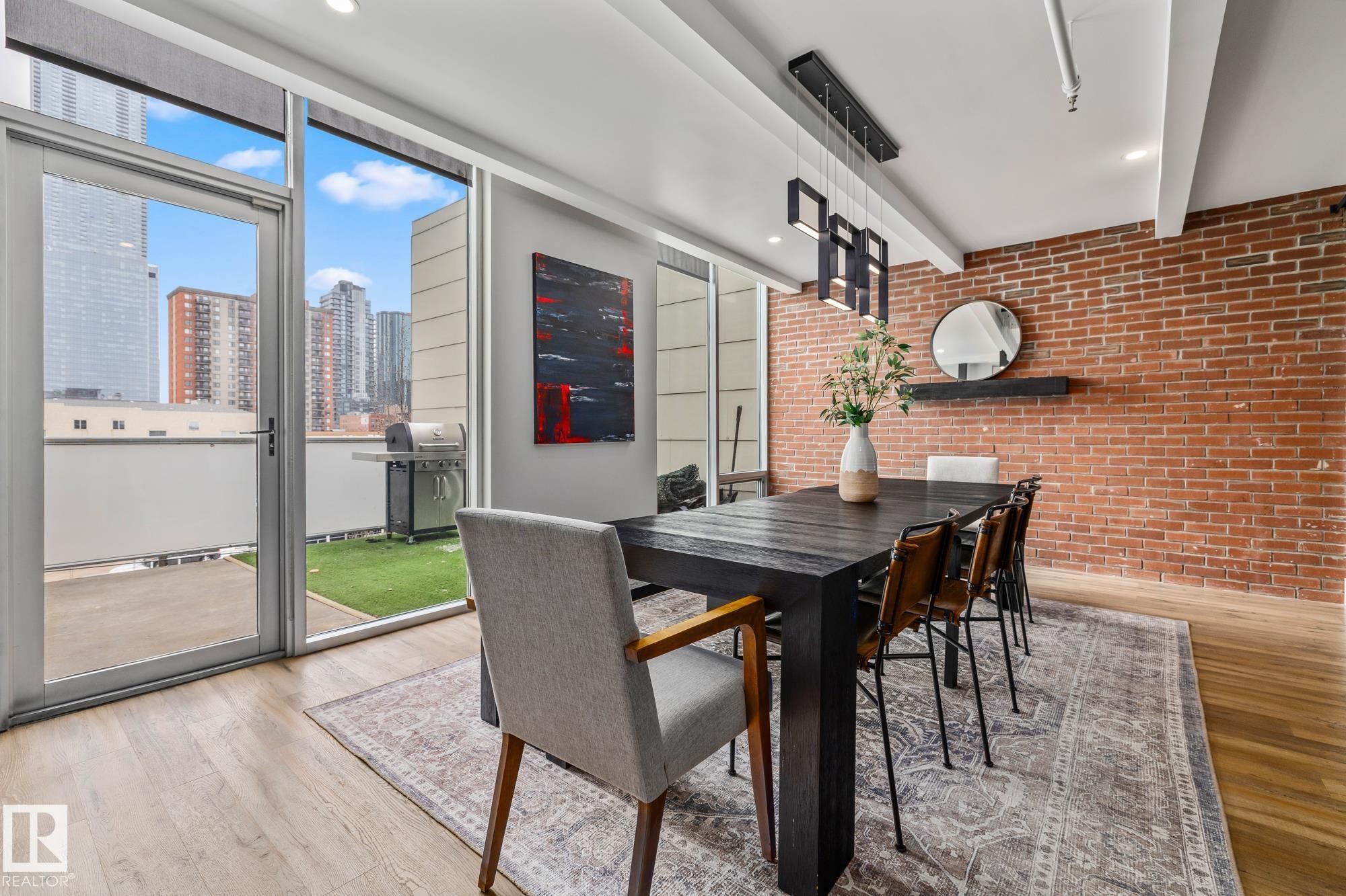 This dining area features an exposed brick accent wall, light wood flooring, and a contemporary chandelier - 314 10309 107 Street, Edmonton, AB -