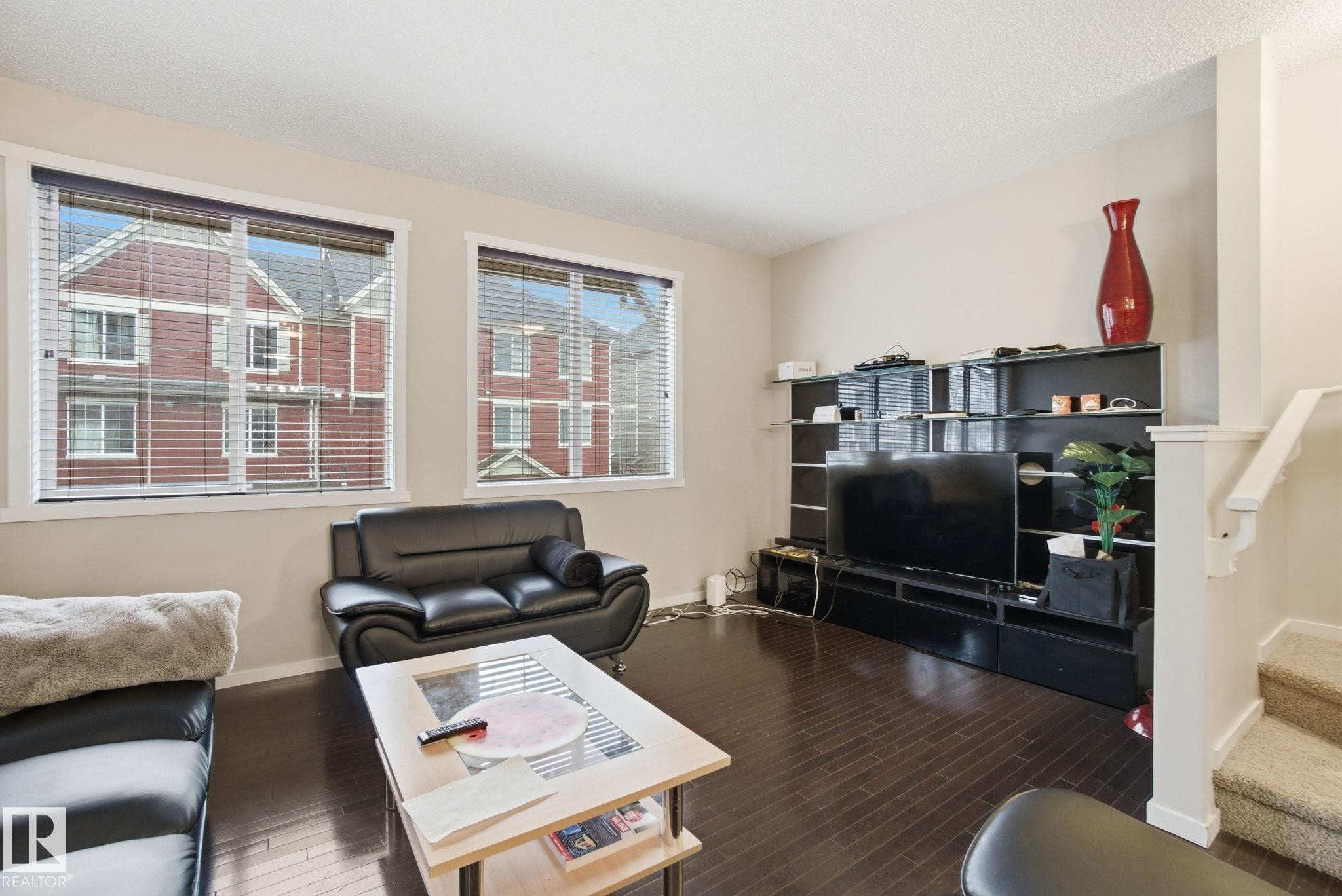 Living area featuring dark hardwood floors, two windows with blinds, and a staircase with carpeting - 75 603 Watt Boulevard, Edmonton, AB - Indoor Photo Showing Living Room