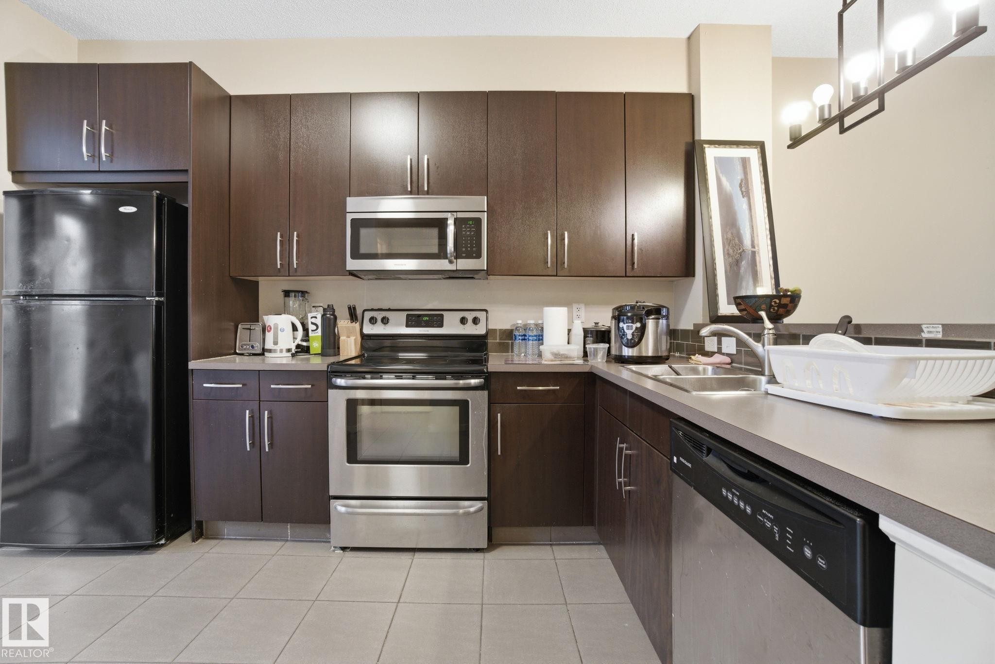 The kitchen features dark wood cabinetry, stainless steel appliances including an oven and microwave, and a black refrigerator - 75 603 Watt Boulevard, Edmonton, AB - Indoor Photo Showing Kitchen With Double Sink