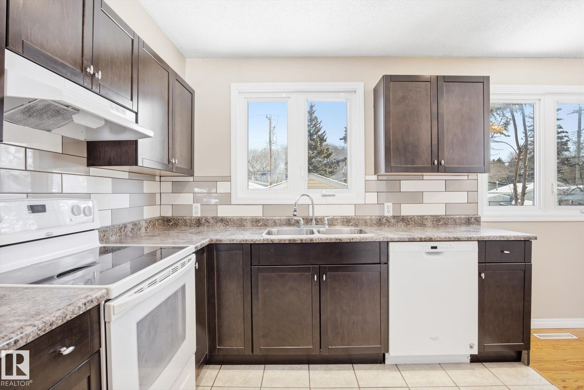 The kitchen features dark wood cabinetry, a subway tile backsplash, a double basin sink, and white appliances - 6 Garden Crescent, St. Albert, AB - Indoor Photo Showing Kitchen With Double Sink