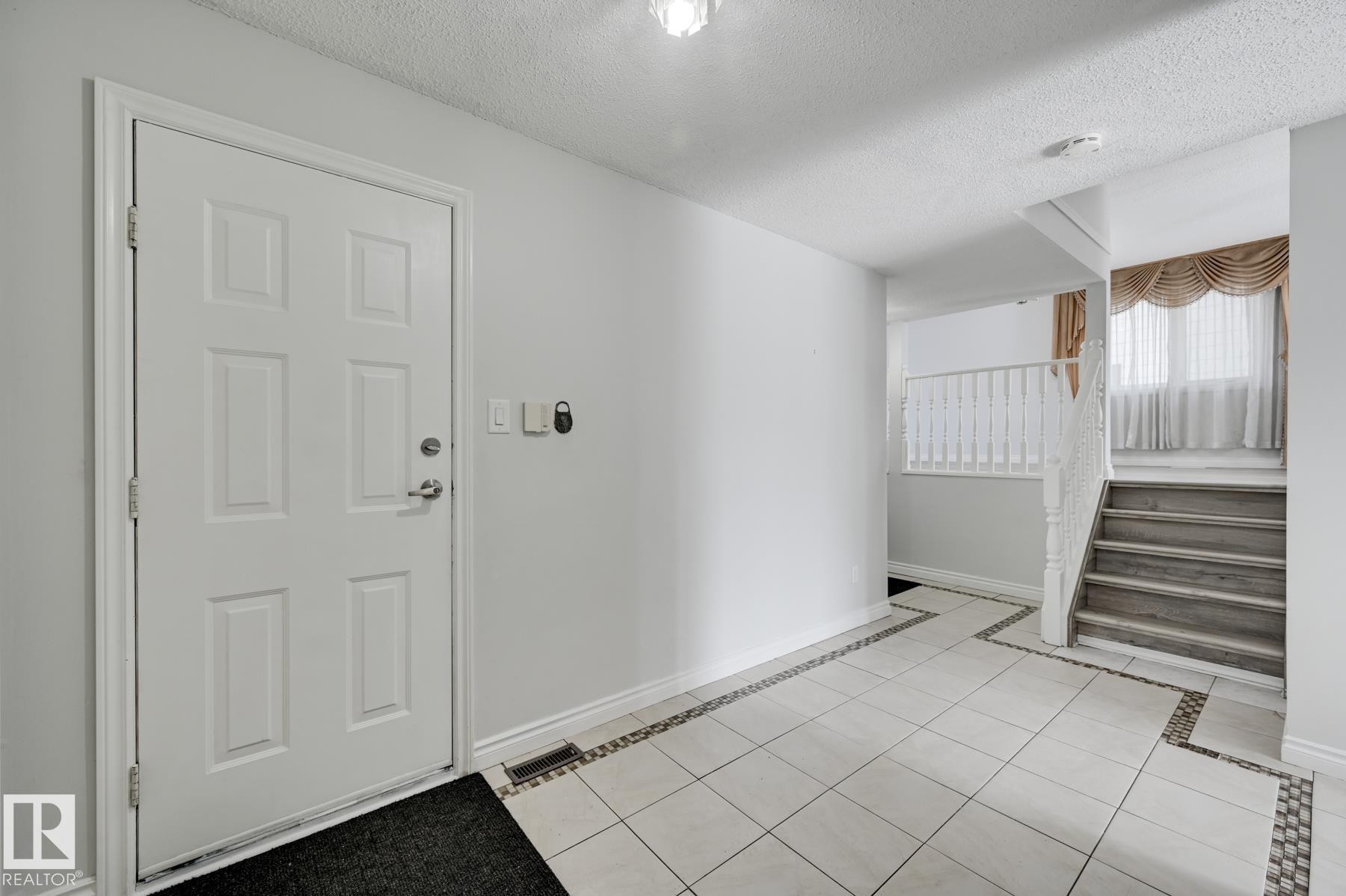 Entryway featuring a white paneled door, light-colored tile flooring with decorative borders, and a staircase with white railings - 8507 152B Avenue, Edmonton, AB - Indoor Photo Showing Other Room