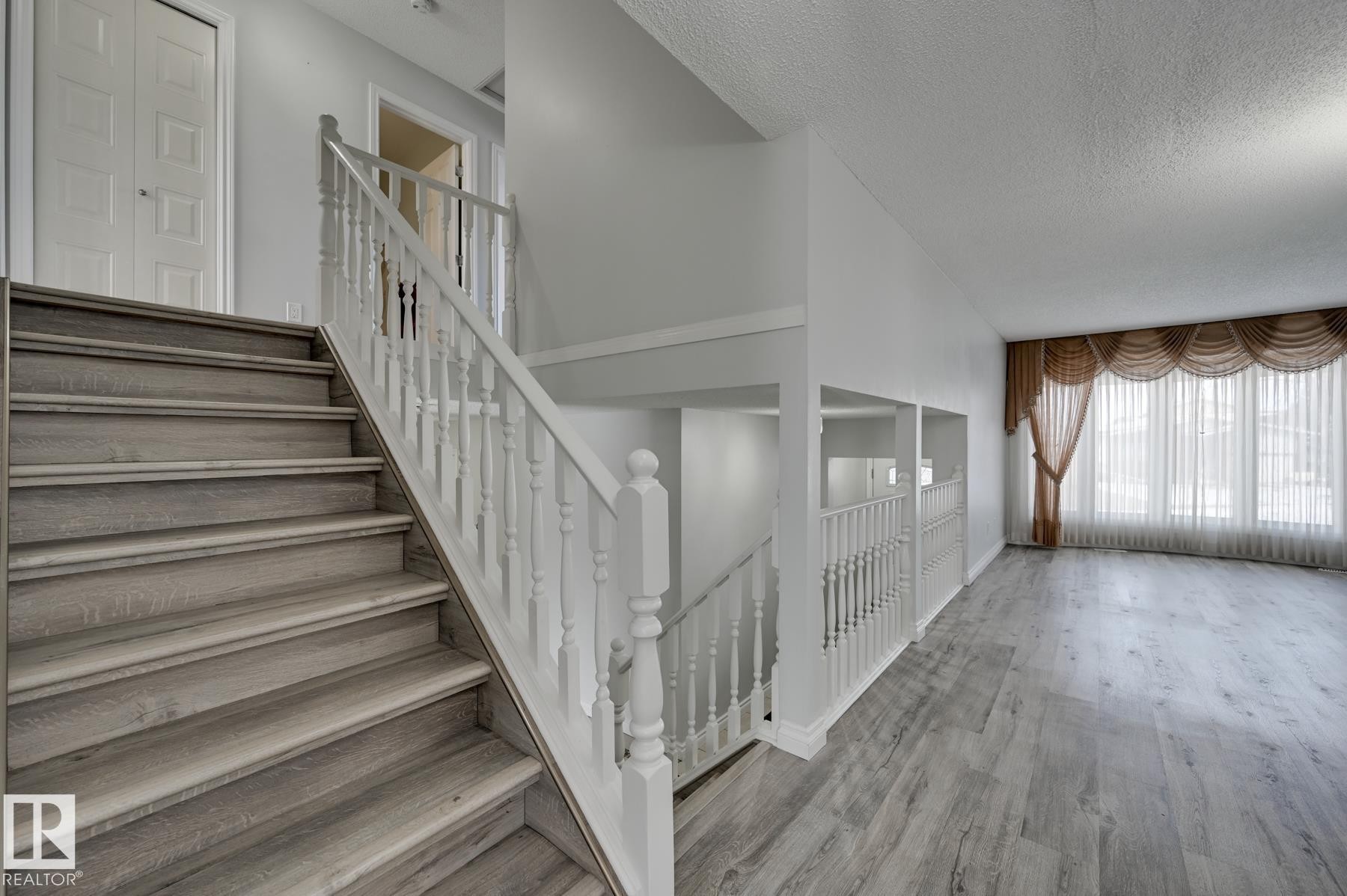 The property features light-toned wood-look flooring throughout, a white staircase with matching balusters, and a spacious living area with large windows - 8507 152B Avenue, Edmonton, AB - Indoor Photo Showing Other Room