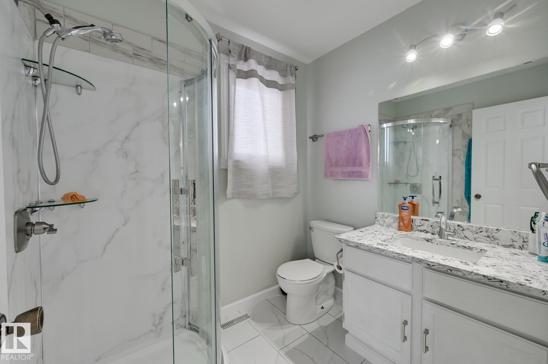 The bathroom features a curved glass shower enclosure with a marble-patterned surround, a white vanity with a patterned countertop, and a mirror with track lighting above - 8507 152B Avenue, Edmonton, AB - Indoor Photo Showing Bathroom