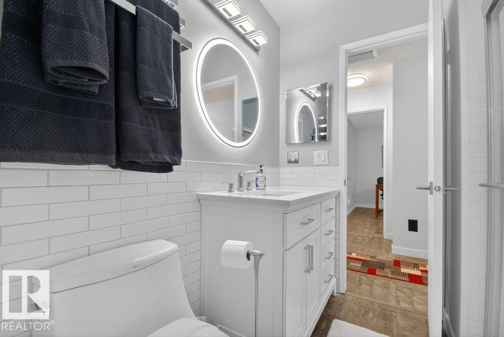 This bathroom features a white subway tile wall, a white vanity with storage, and a round, illuminated mirror - 91 Hearthstone, Edmonton, AB - Indoor Photo Showing Bathroom