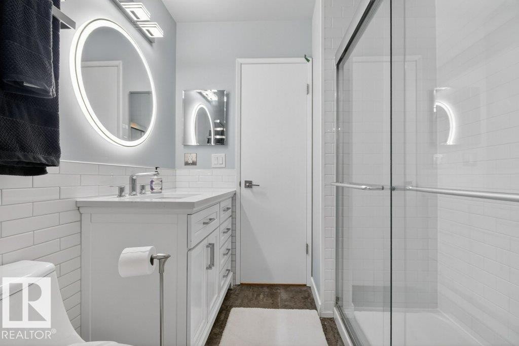 Bathroom featuring a white subway tile accent wall, a modern vanity with a white countertop, an illuminated round mirror, and a glass-enclosed shower - 91 Hearthstone, Edmonton, AB - Indoor Photo Showing Bathroom