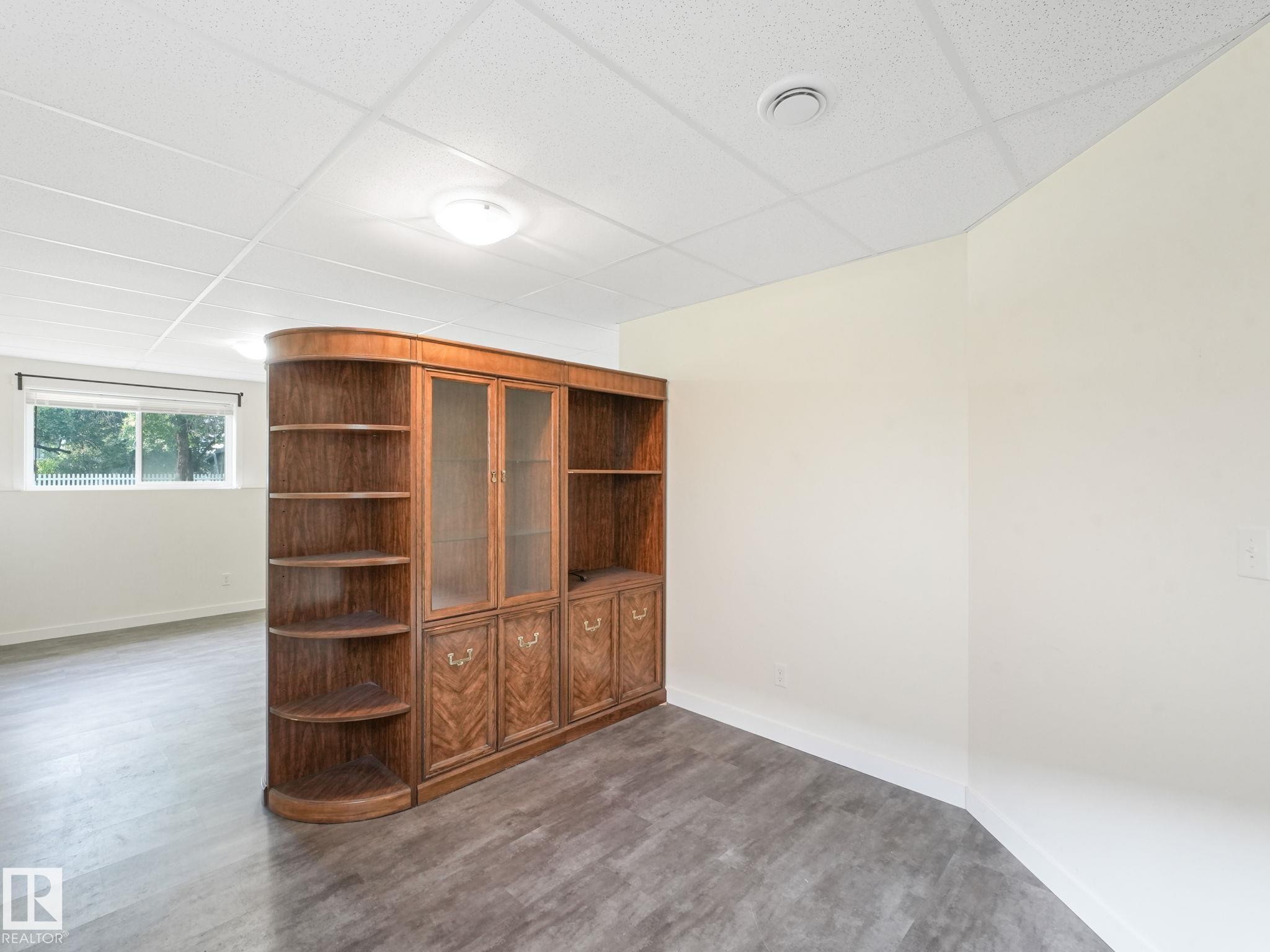 This room features a built-in wooden cabinet with glass doors and shelving, light-colored walls, and a window providing natural light - 5035 51 Avenue, Lamont, AB - Indoor Photo Showing Other Room
