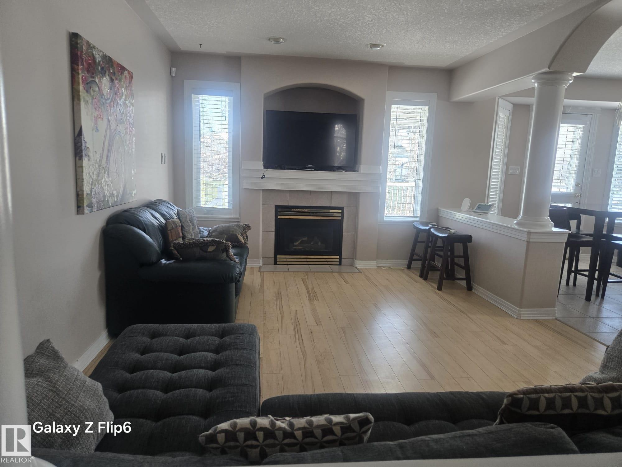 This living area features light-toned hardwood flooring, a fireplace with a mantel, and windows providing natural light - Edmonton, AB - Indoor Photo Showing Living Room With Fireplace