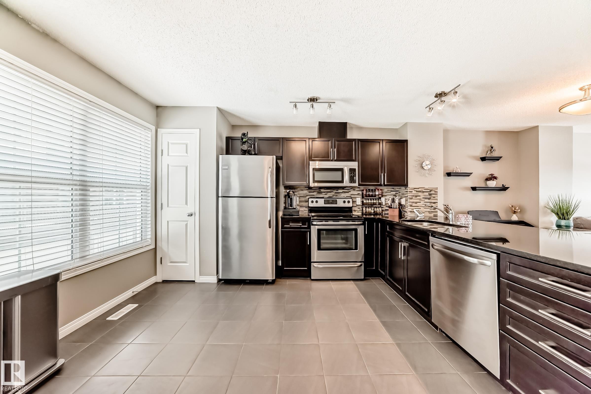 The kitchen features dark wood cabinetry, stainless steel appliances, a tiled backsplash, and dark tiled flooring - 86 4029 Orchards Drive, Edmonton, AB - Indoor Photo Showing Kitchen