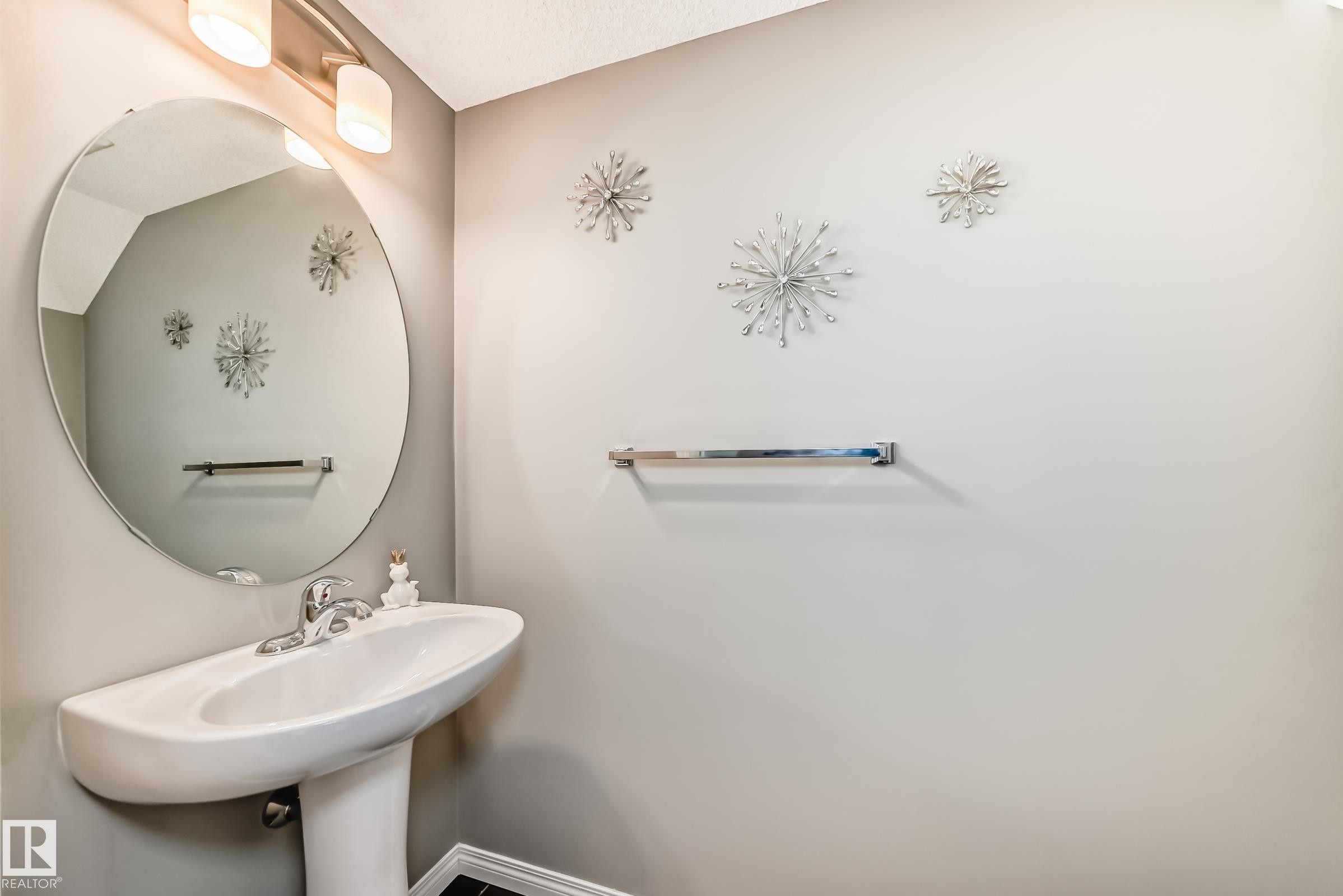 The property features a vanity with a white pedestal sink, a chrome faucet, and a round mirror with overhead lighting - 86 4029 Orchards Drive, Edmonton, AB - Indoor Photo Showing Bathroom