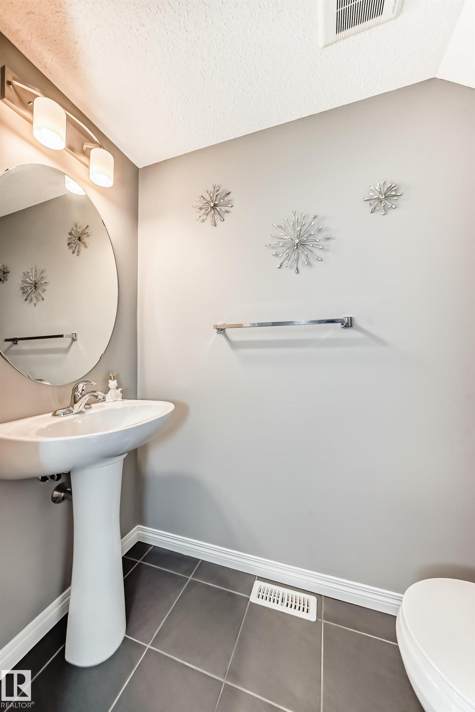The bathroom features a pedestal sink with a chrome faucet, a round mirror, and a wall-mounted light fixture with three sconces - 86 4029 Orchards Drive, Edmonton, AB - Indoor Photo Showing Bathroom