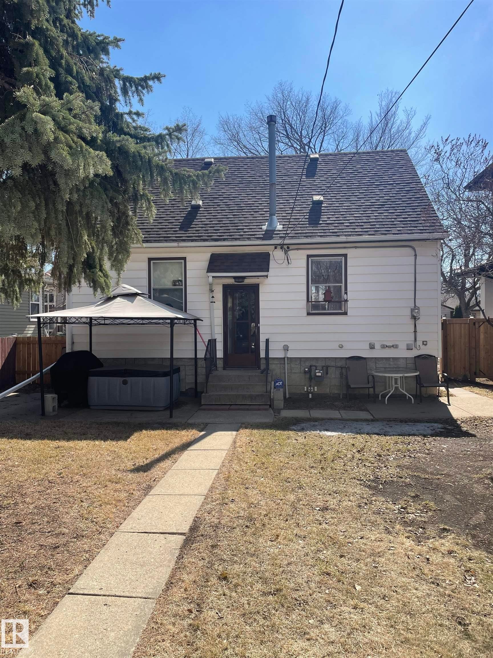 The property features a white exterior, a dark shingled roof, and a mature tree providing shade - 7934 81 Avenue, Edmonton, AB - Outdoor