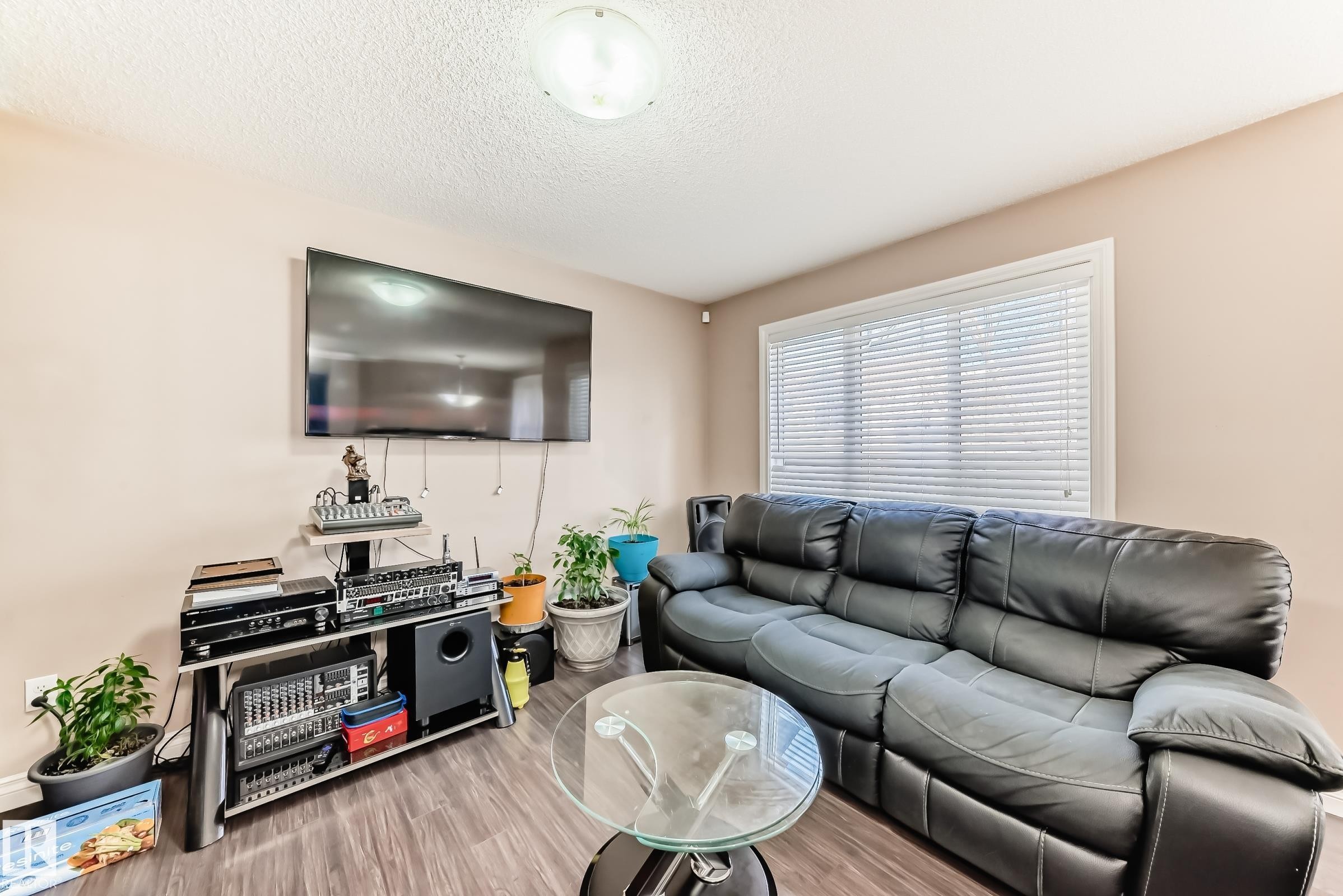 The living area features a window with horizontal blinds, wood-look flooring, and a ceiling light fixture - 23 445 Brintnell Boulevard Nw, Edmonton, AB - Indoor Photo Showing Living Room