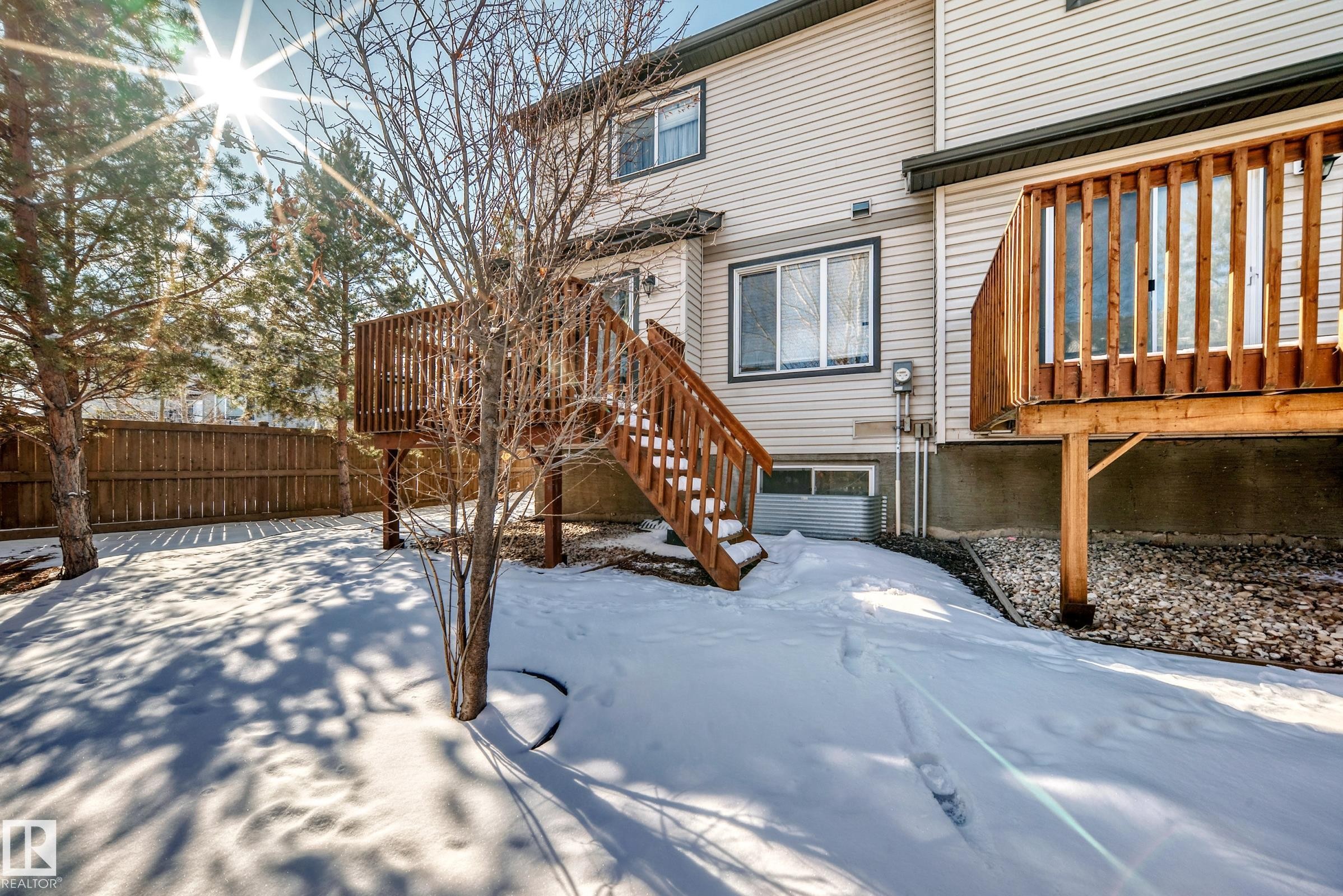 The property features a wooden privacy fence, mature trees, and a wooden deck with stairs leading to the yard - 23 445 Brintnell Boulevard Nw, Edmonton, AB - Outdoor