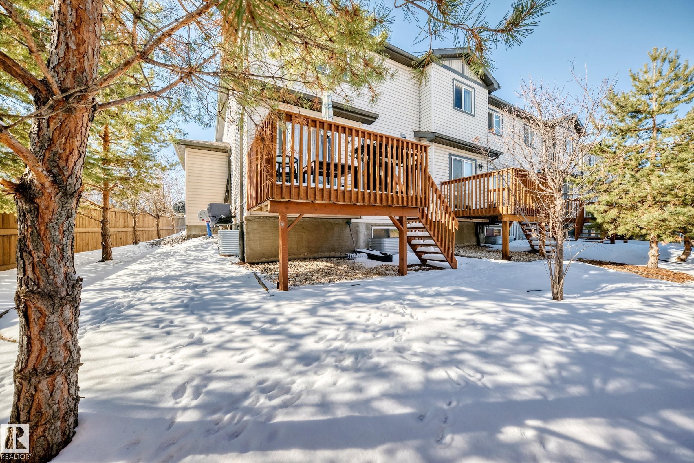 The property features a private yard with extensive snow cover and mature trees, alongside two wooden decks with railings and stairs leading into the yard - 23 445 Brintnell Boulevard Nw, Edmonton, AB - Outdoor With Deck Patio Veranda