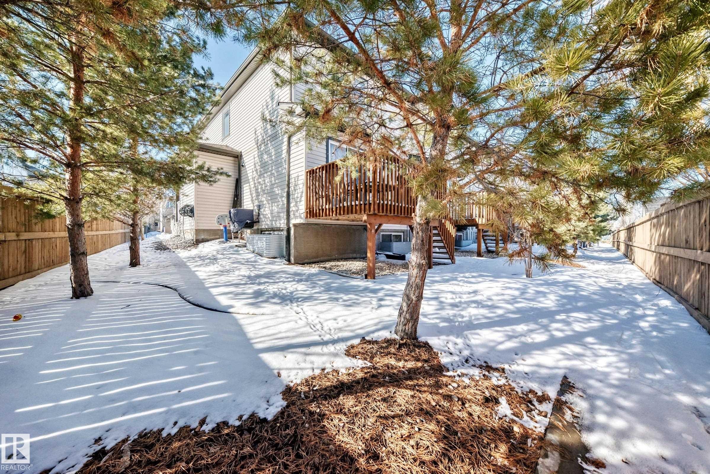 The property features a fenced yard with mature trees and a wooden deck, accessible via stairs - 23 445 Brintnell Boulevard Nw, Edmonton, AB - Outdoor