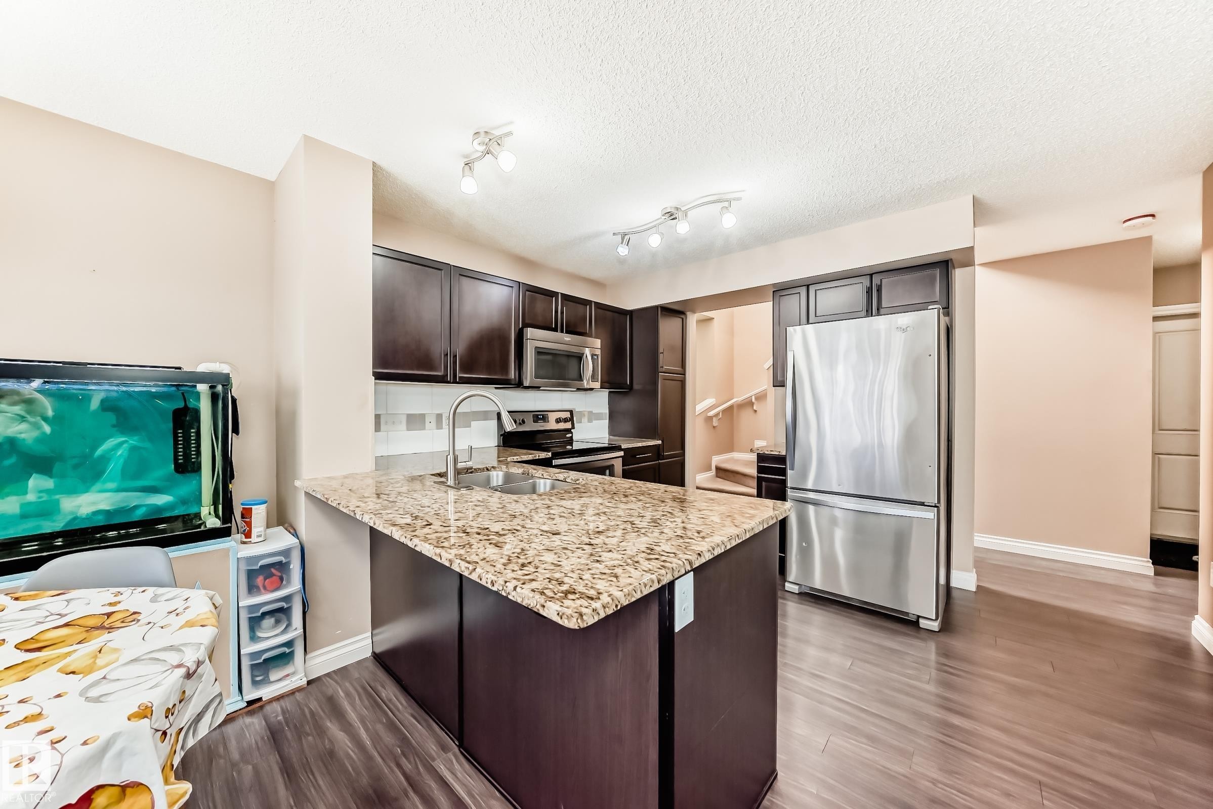 Well-appointed kitchen featuring dark wood cabinetry, a stainless steel refrigerator, and a kitchen island with a granite countertop and sink - 23 445 Brintnell Boulevard Nw, Edmonton, AB - Indoor Photo Showing Kitchen With Double Sink