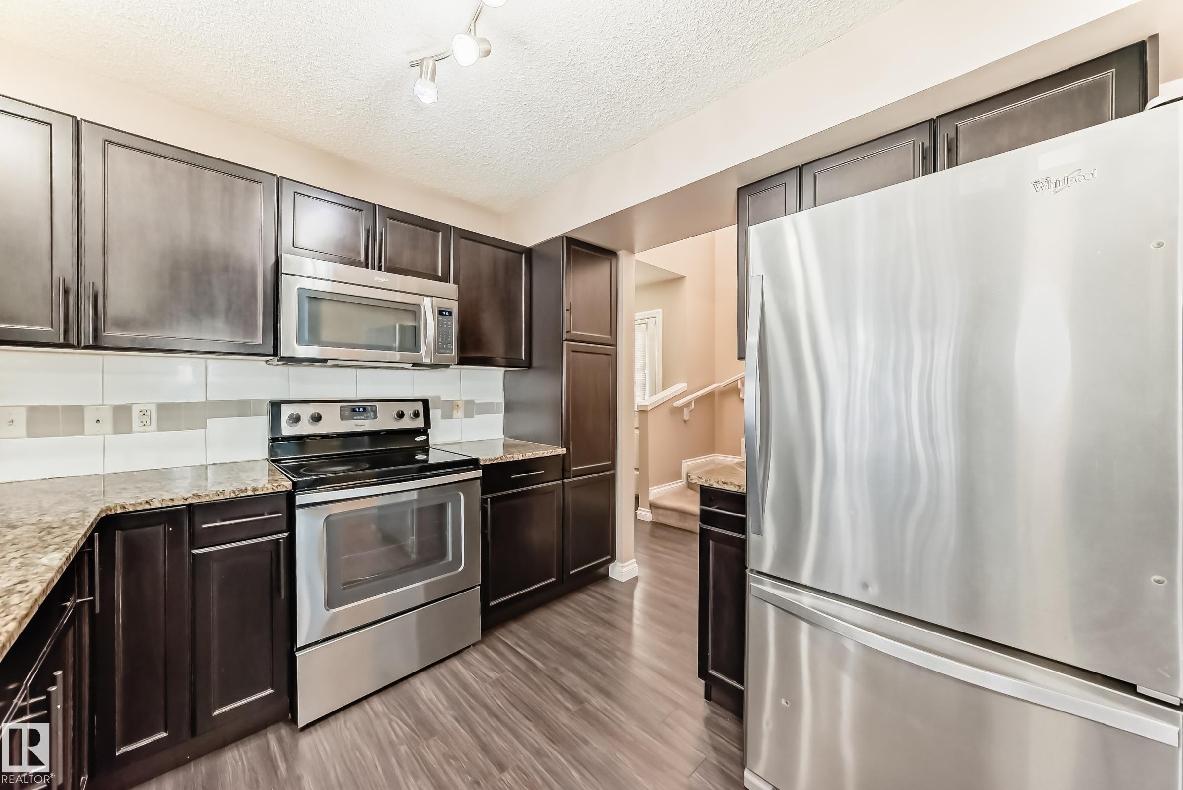 The kitchen features dark wood cabinetry, granite countertops, and stainless steel appliances including a refrigerator, stove, and microwave - 23 445 Brintnell Boulevard Nw, Edmonton, AB - Indoor Photo Showing Kitchen