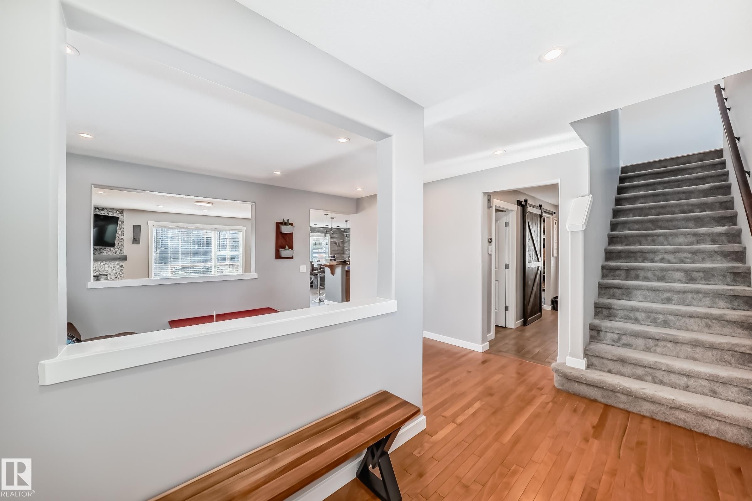 The property features hardwood flooring in the entry, a carpeted staircase with a dark wood handrail, and a pass-through window looking into a room with a large window - 6303 2 Avenue, Edmonton, AB - Indoor Photo Showing Other Room