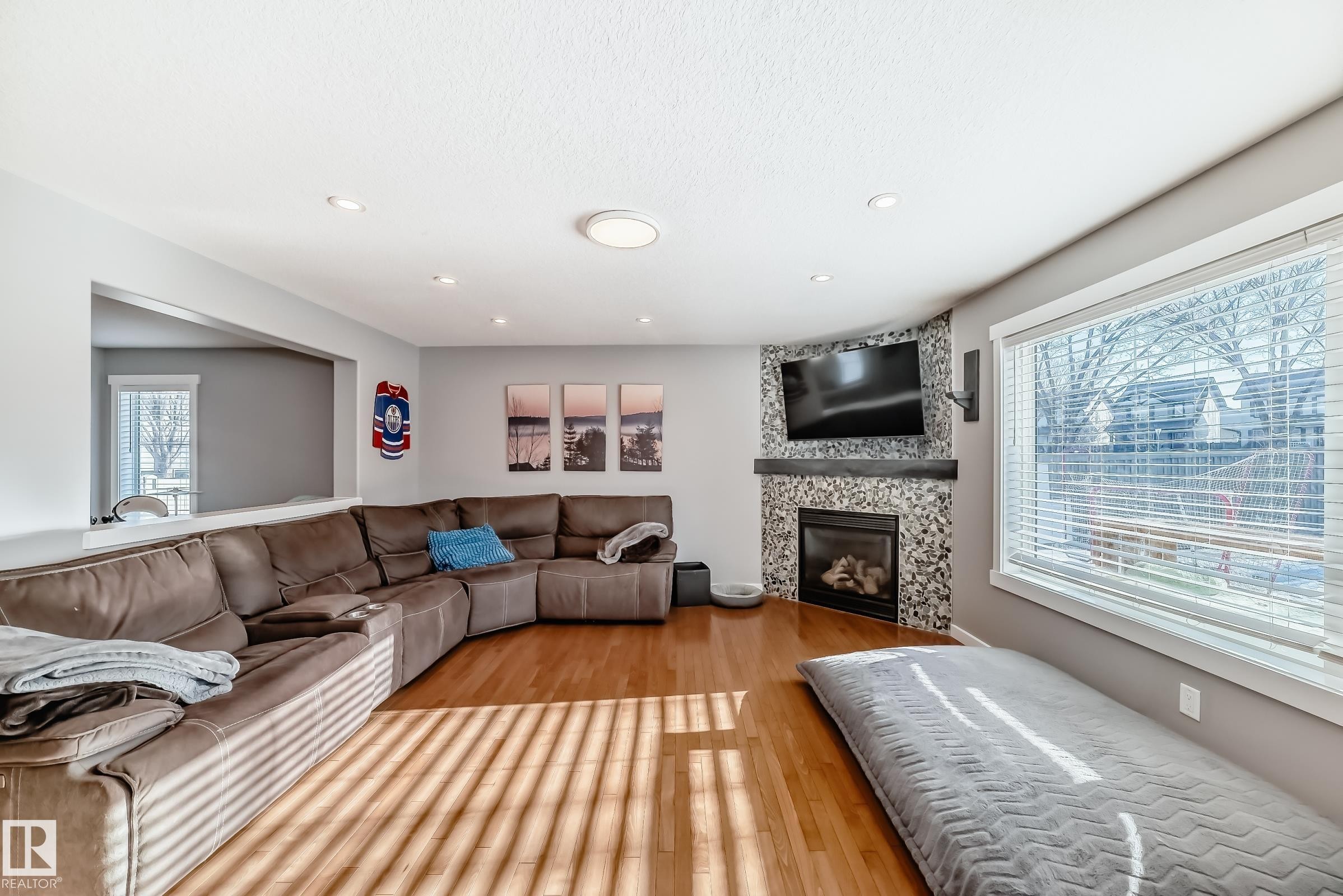 Living room featuring hardwood flooring, a fireplace with a stone surround and wood mantel, and large windows with blinds - 6303 2 Avenue, Edmonton, AB - Indoor Photo Showing Living Room With Fireplace