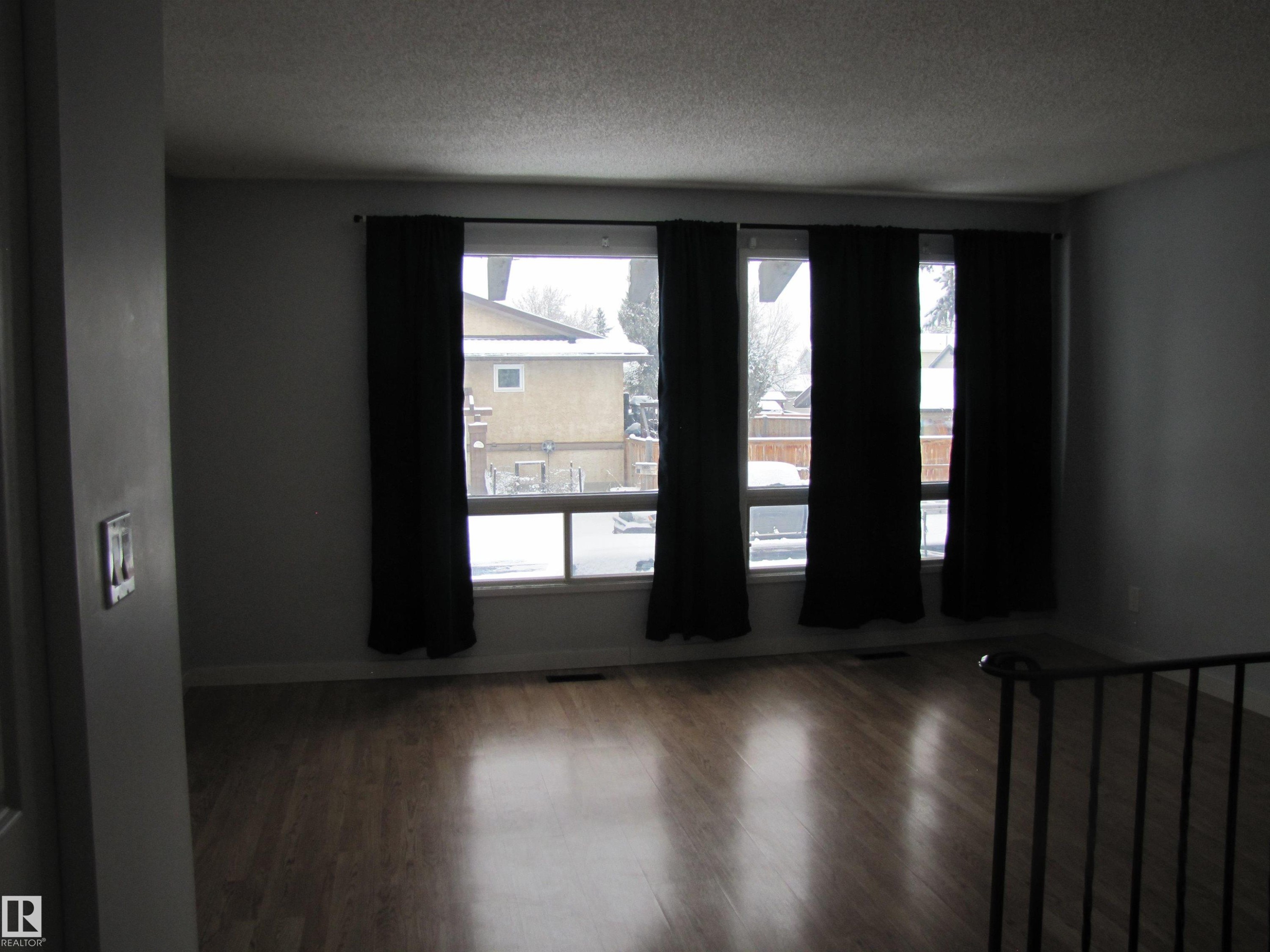 This room features large windows with dark curtains, warm-toned wood flooring, and light-colored walls - 3617 43A Avenue, Edmonton, AB - Indoor Photo Showing Other Room