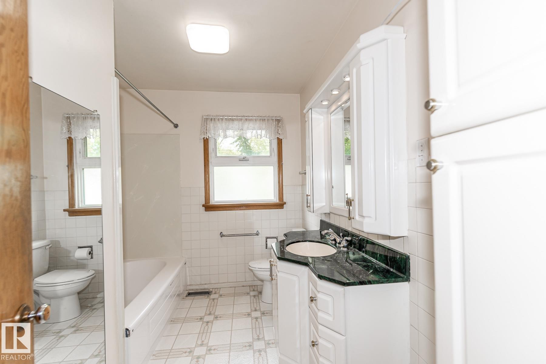 The bathroom features a vanity with a dark green countertop, white cabinetry, a bathtub, and white tiled flooring - 11815 76 Avenue, Edmonton, AB - Indoor Photo Showing Bathroom