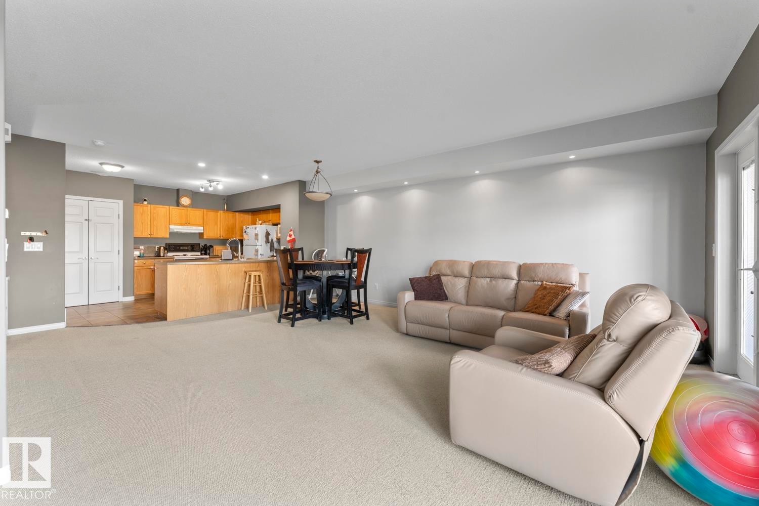 Open concept living area with light-colored carpeting, recessed lighting, and a view of the kitchen featuring wooden cabinetry and stainless steel appliances - 107 801 11 Street, Cold Lake, AB - Indoor