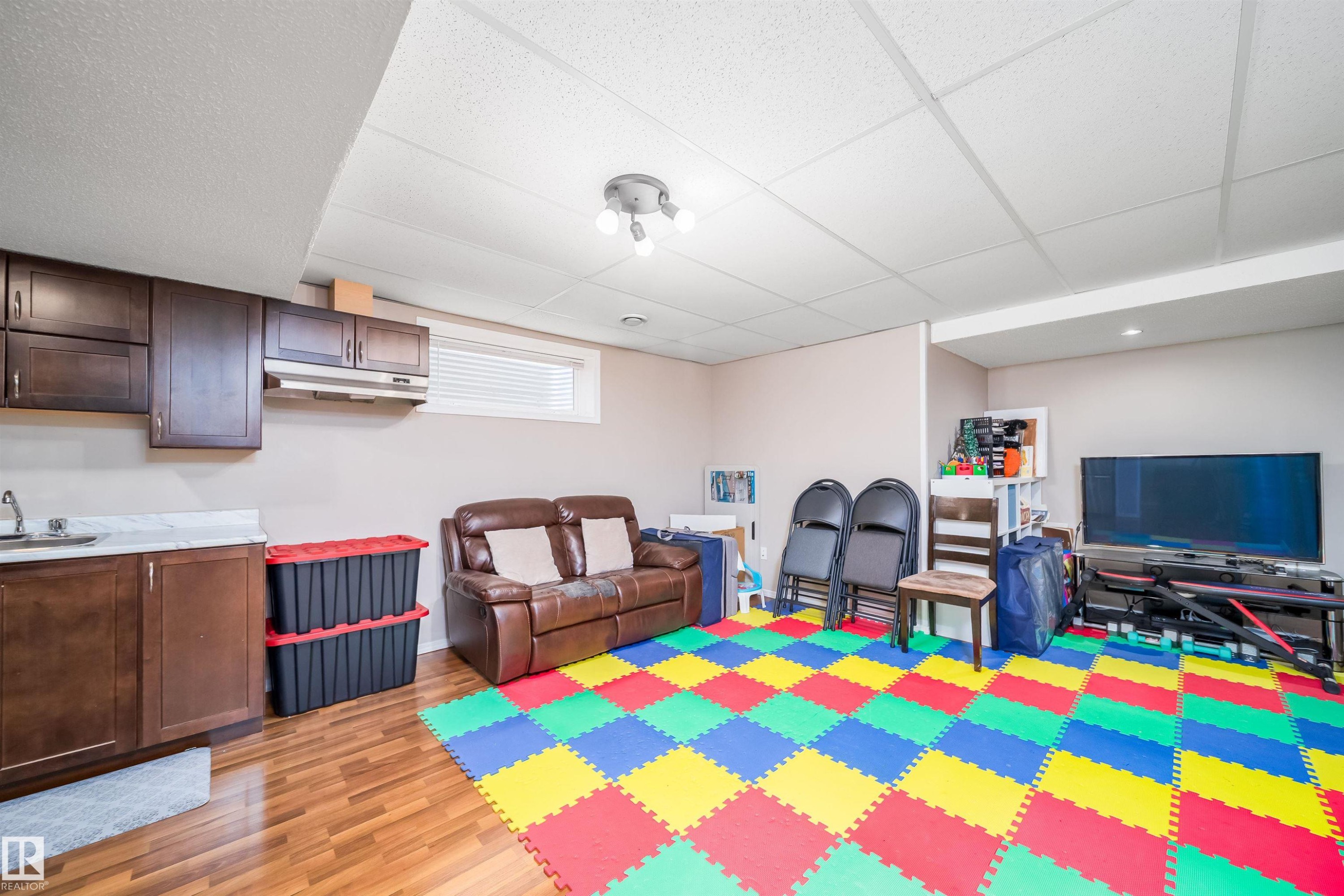 Spacious room featuring wood-look flooring, a drop ceiling, and a functional wet bar with wood cabinetry and a light-colored countertop - 8932 175 Avenue, Edmonton, AB - Indoor