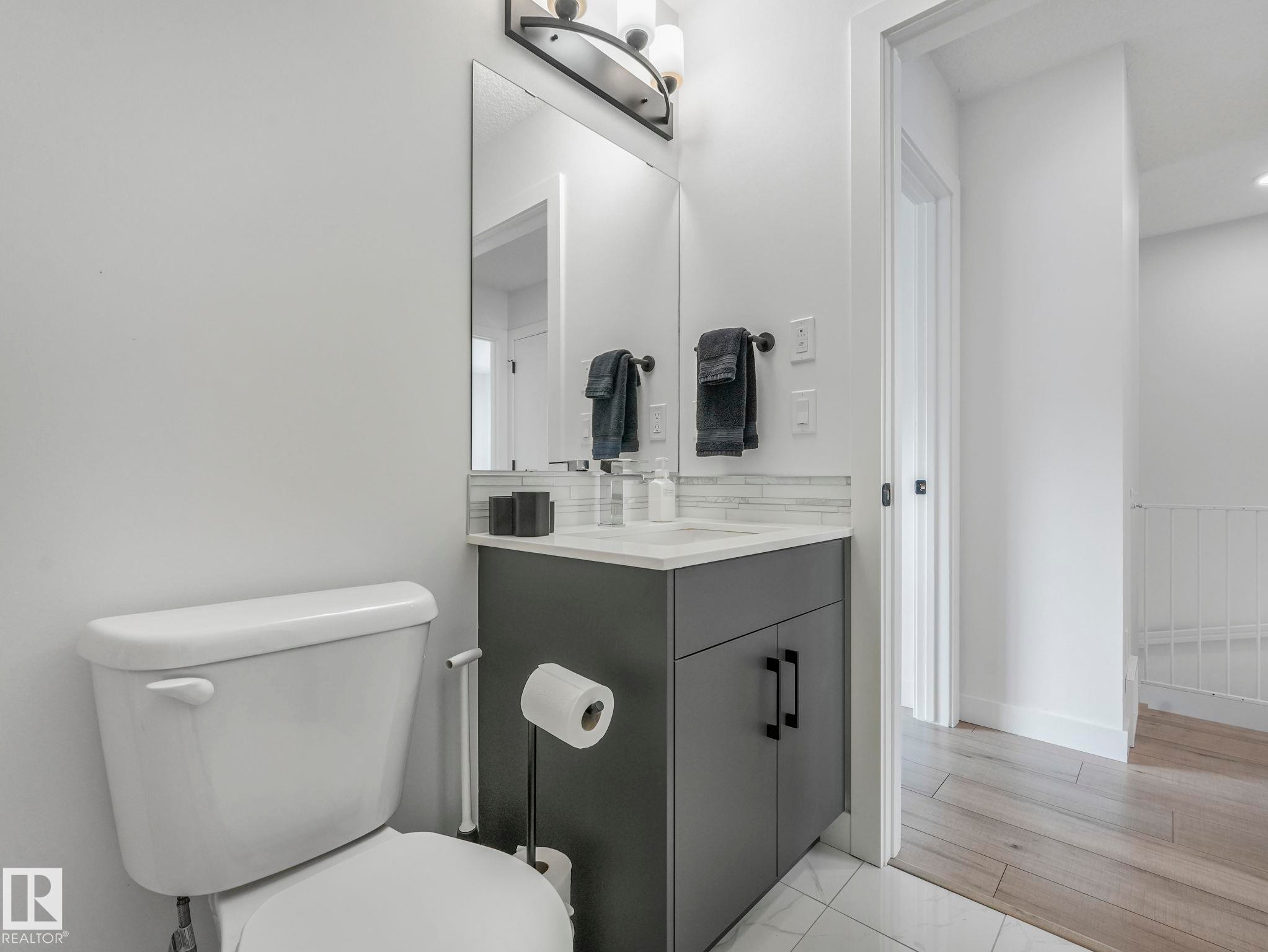 Bathroom featuring a white toilet, a vanity with a dark grey cabinet and white countertop, a mirror, and a wall-mounted light fixture - 95 1010 Rabbit Hill Road, Edmonton, AB - Indoor Photo Showing Bathroom
